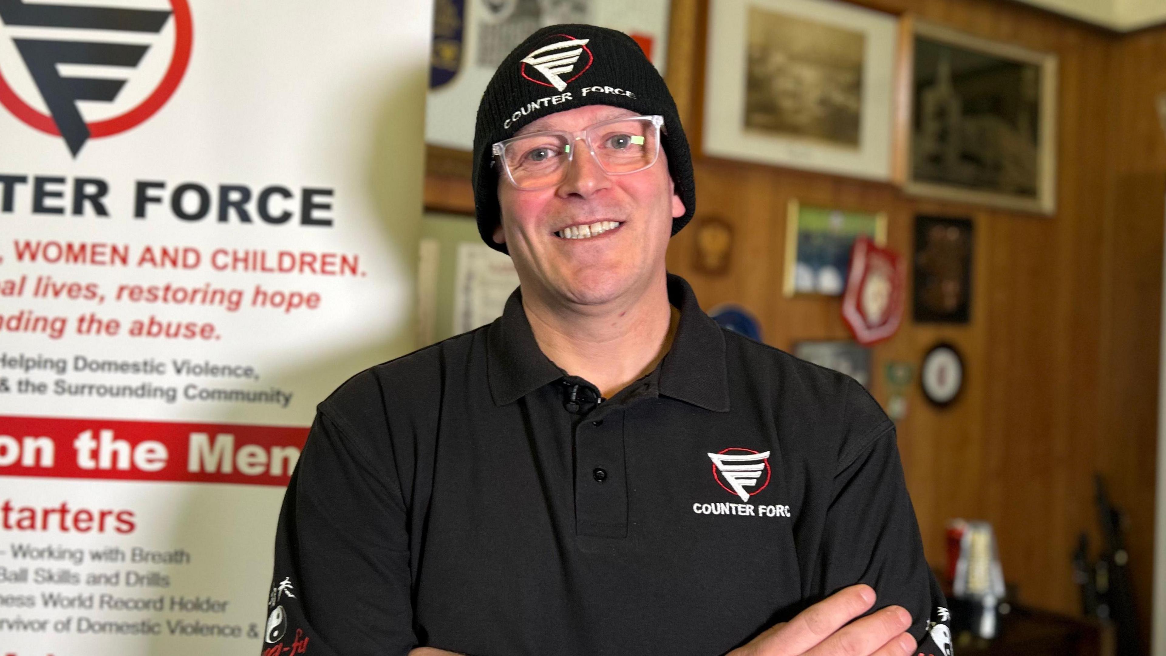 Carl Fitzpatrick yn gwisgo het ddu gyda logo Counter Force a chrys-t du gyda logo Counter Force. Yn gwisgo sbectol ac yn croesi ei freichiau.
