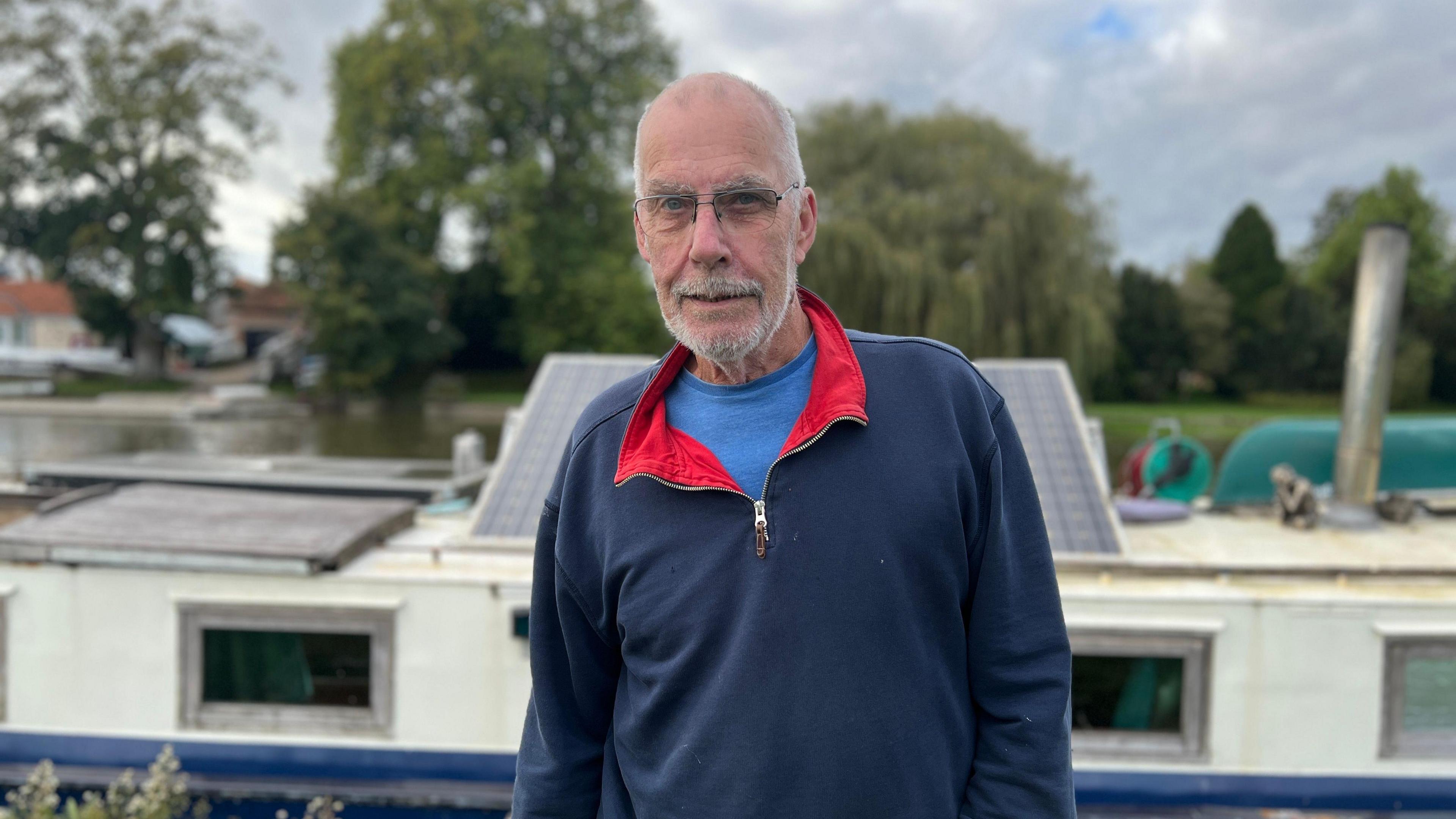 A grey-haired man wearing a blue sweatshirt looking straight at the camera. The roof of his boat and the river are in soft focus behind him.