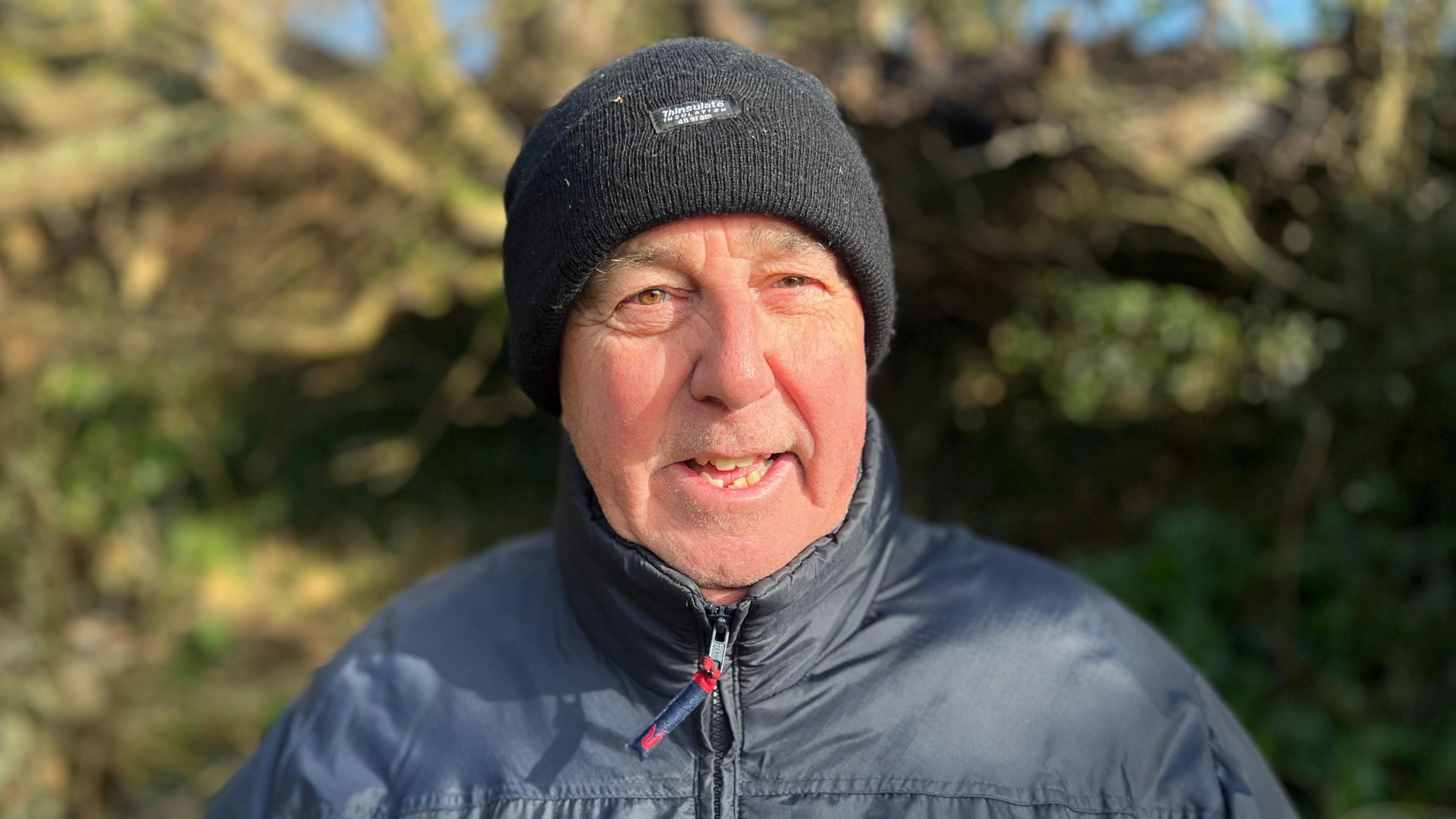 Mike Chantry wearing a black woolly hat with a navy blue waterproof coat in front of a fallen tree