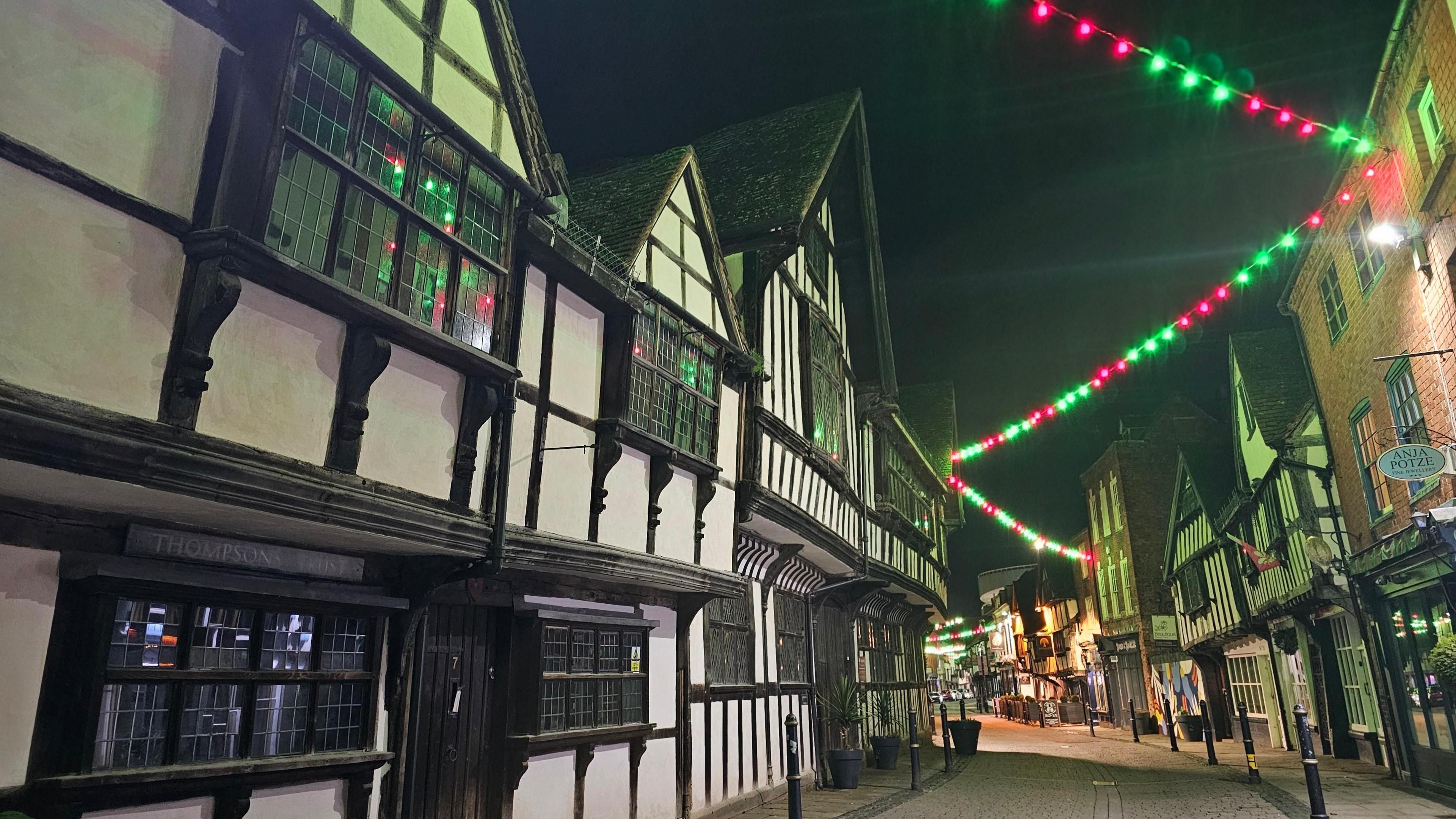 Black-and-white half-timbered buildings on both sides of a street with green and pink lights strung between them.