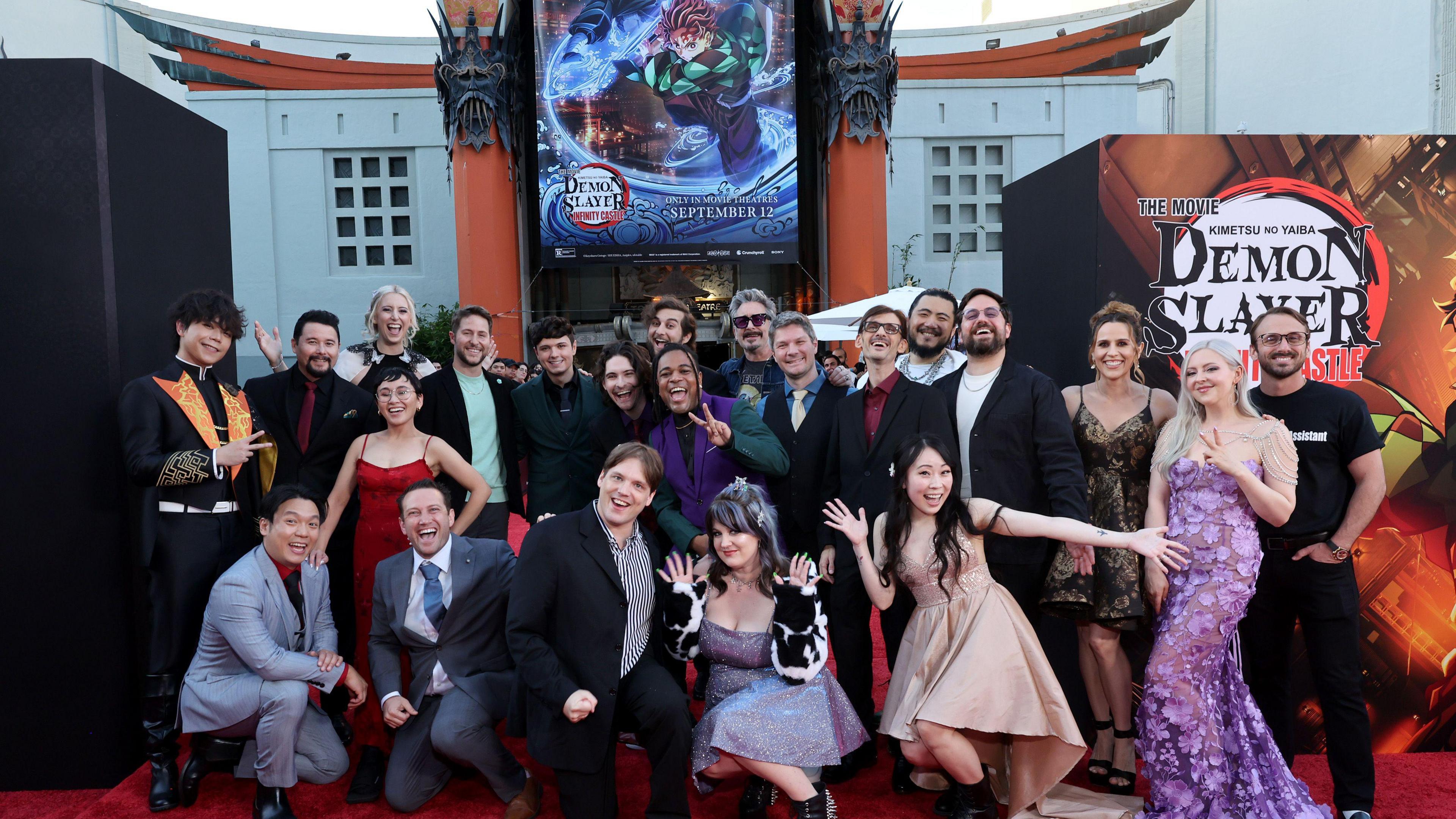 A group of people dressed in suits and gowns gather outside a cinema on a red carpet, flanked by posters for Demon Slayer, Kimetsu no Yaiba Infinity Castle. The mood is celebratory.