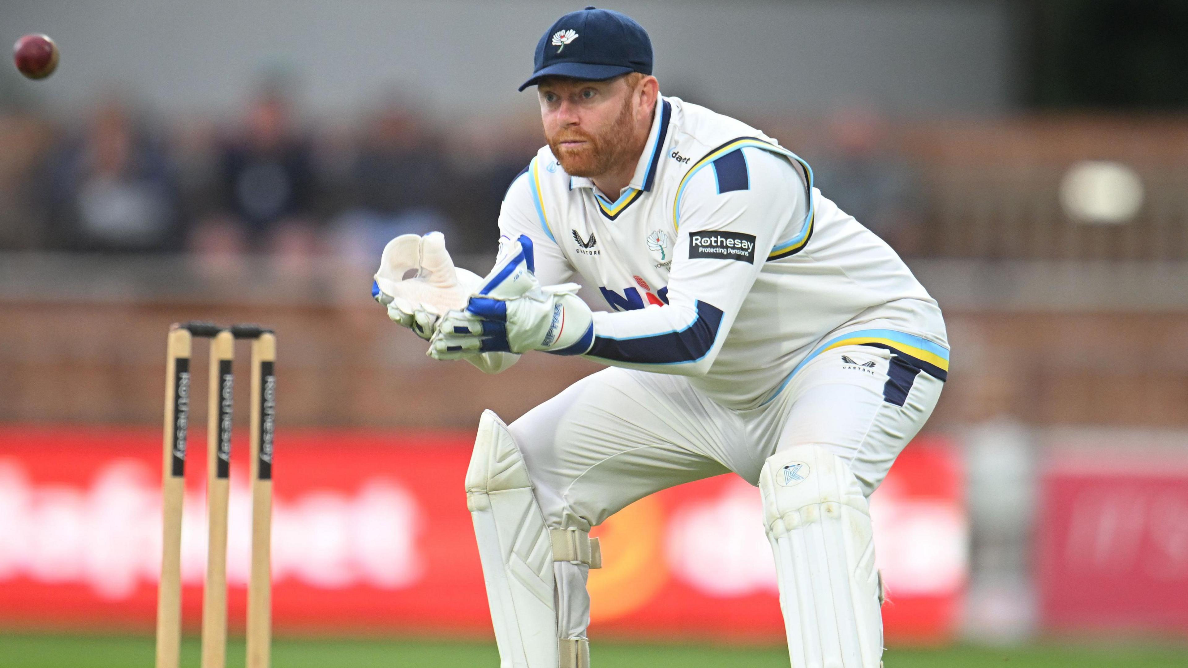 Jonny Bairstow, wearing a white Yorkshire jumper vest and whites with wicket-keeping gloves, pads and a blue hat with a Yorkshire logo on, prepares to catch an incoming ball while wicket-keeping for Yorkshire in 2025 with a look of concentration on his face