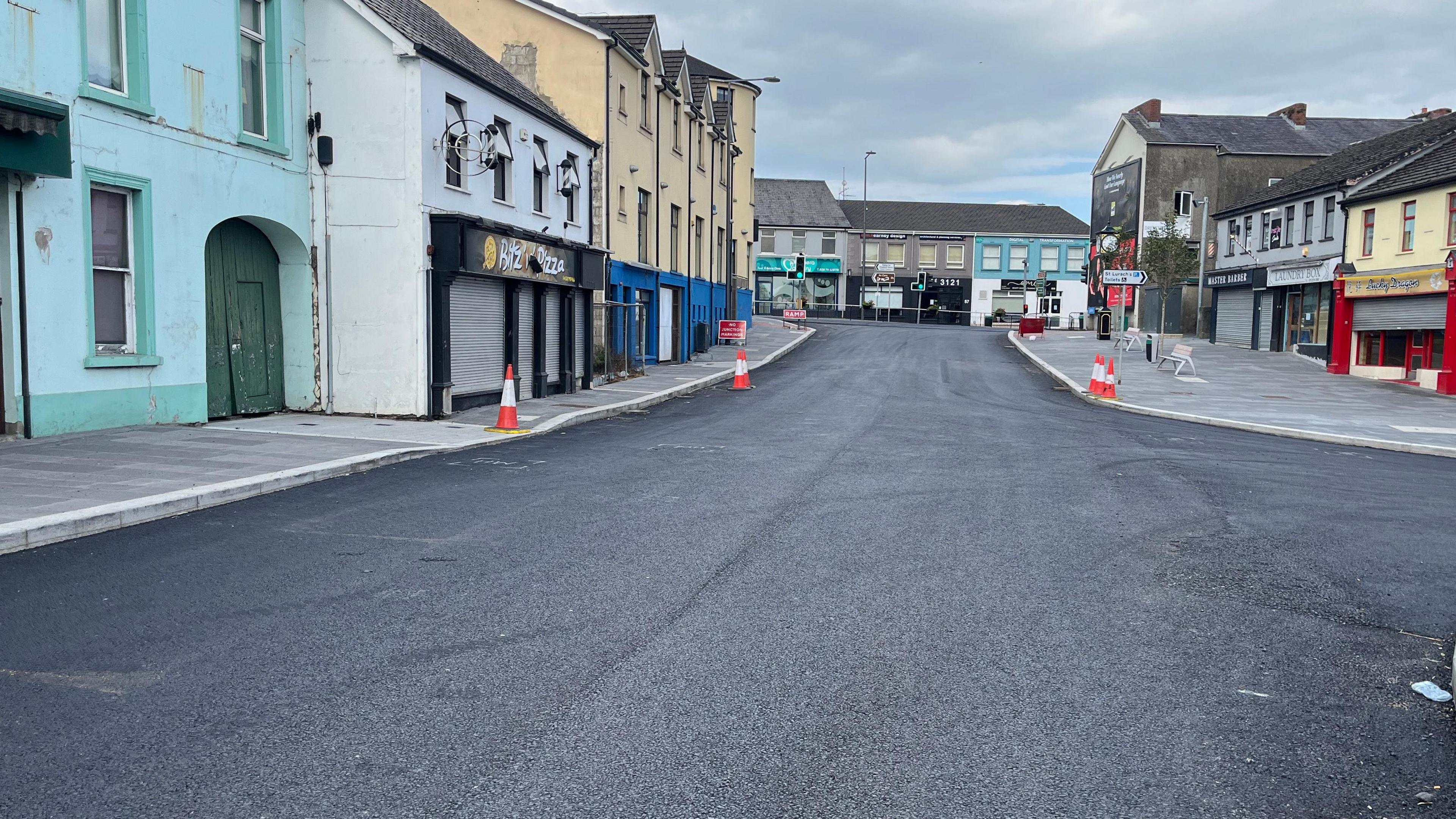 A newly surfaced black tarmac road, with shops and properties on either side. There are a few cones and road signs in the picture.