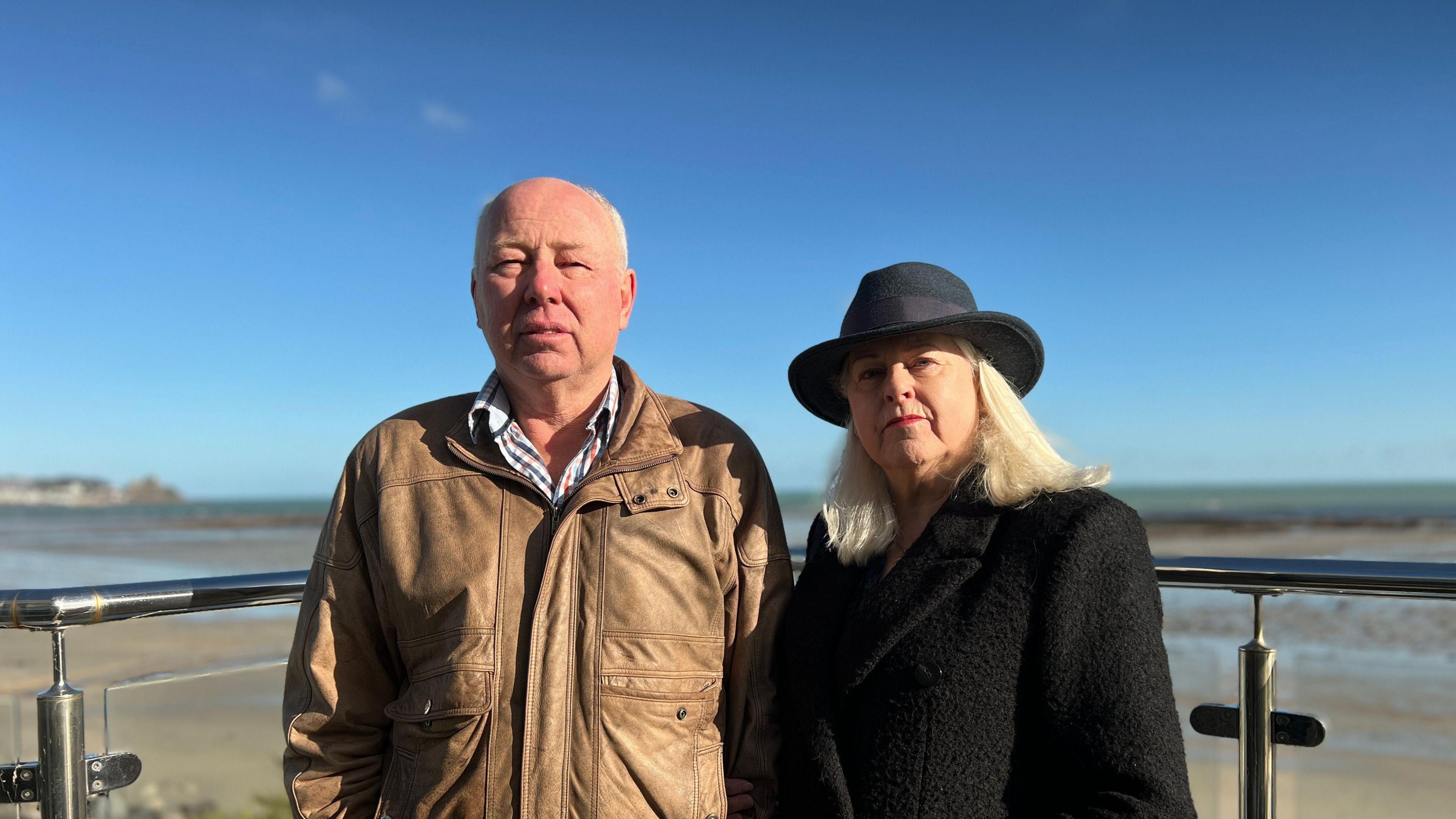 Ted Egre wearing a brown leather jacket and a blue, red and white checked shirt. Jackie Hilton-Egre wearing a black coat with a black hat. They are stood on a balcony overlooking the sea.