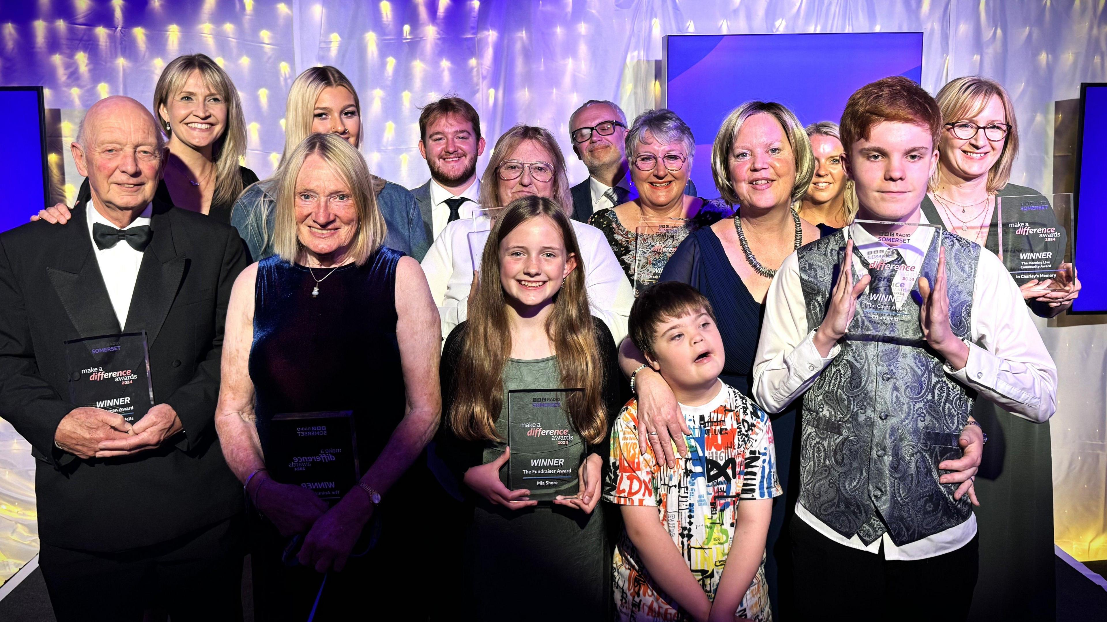 A group image of the winners of the Make A Difference Awards. Everyone is holding a glass rectangular trophy and standing against a white backdrop with fairy lights and a blue hue. 