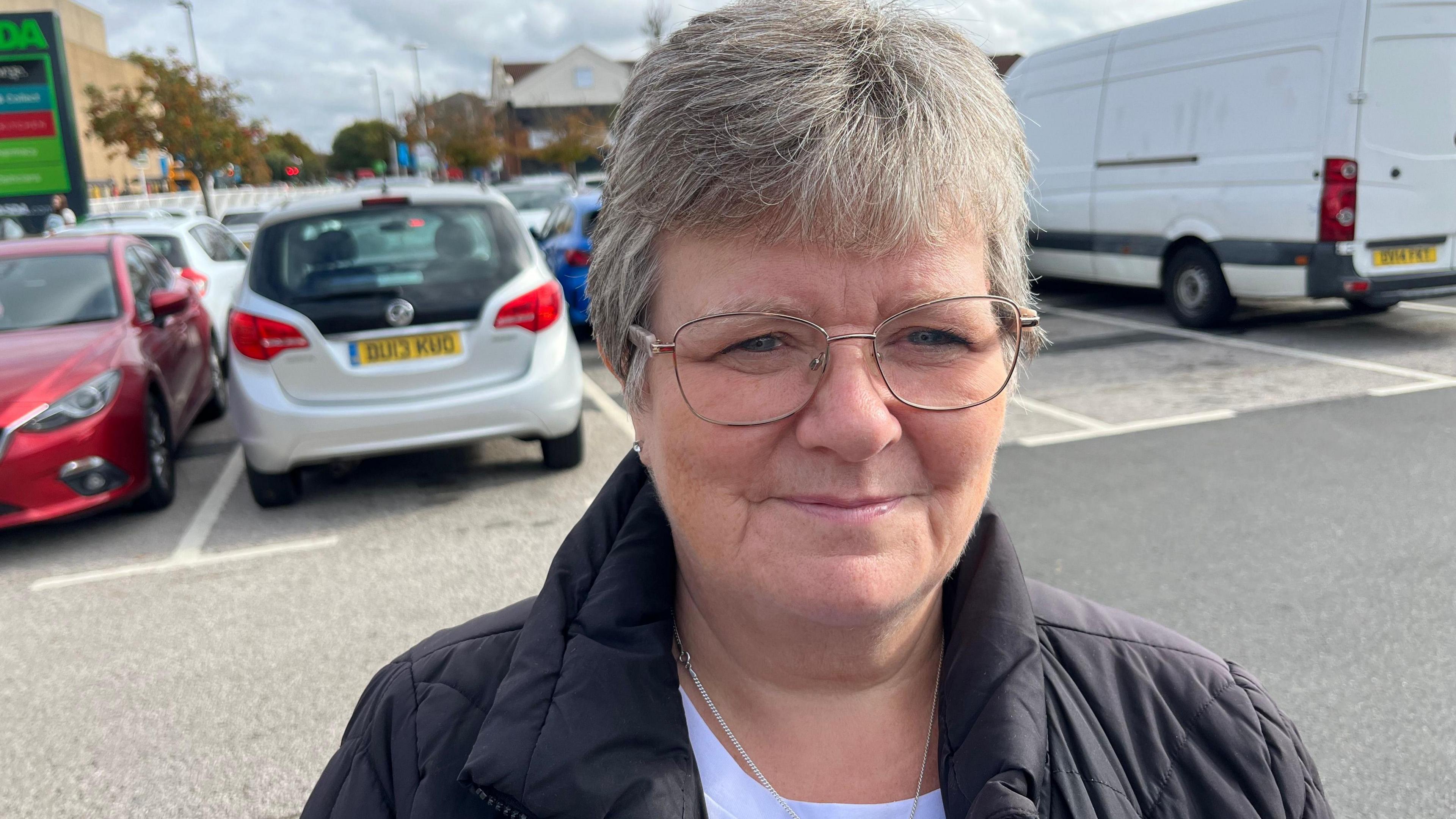 Carolyne Childs smiling for the camera, while standing in an outdoor car park. She has grey hair and glasses and is wearing a white T-shirt under a puffer jacket.