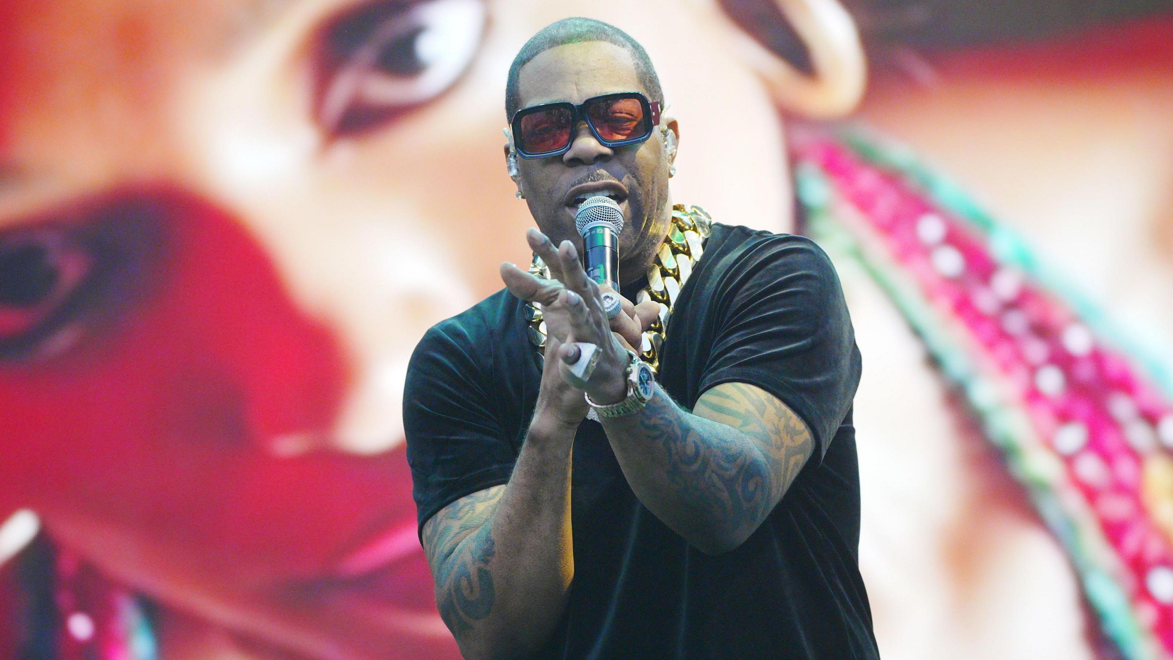 A man wearing a black T-shirt and a big gold chain is holding a microphone on a stage with a picture of a woman on a screen behind him.