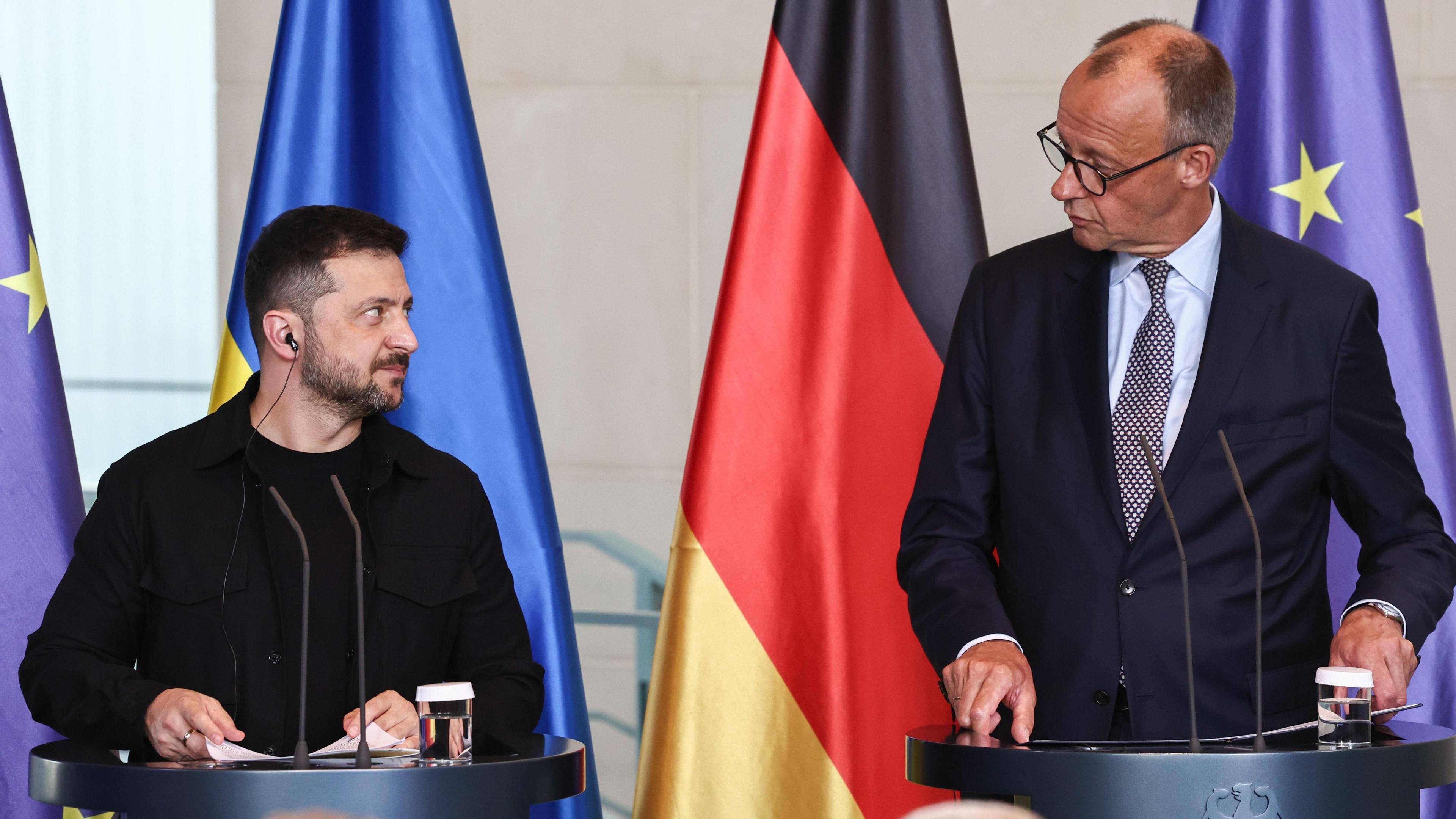 Volodymyr Zelensky and the German chancellor stand in front of two lecterns looking at each other. The flags of Ukraine and Germany are hanging behind them.