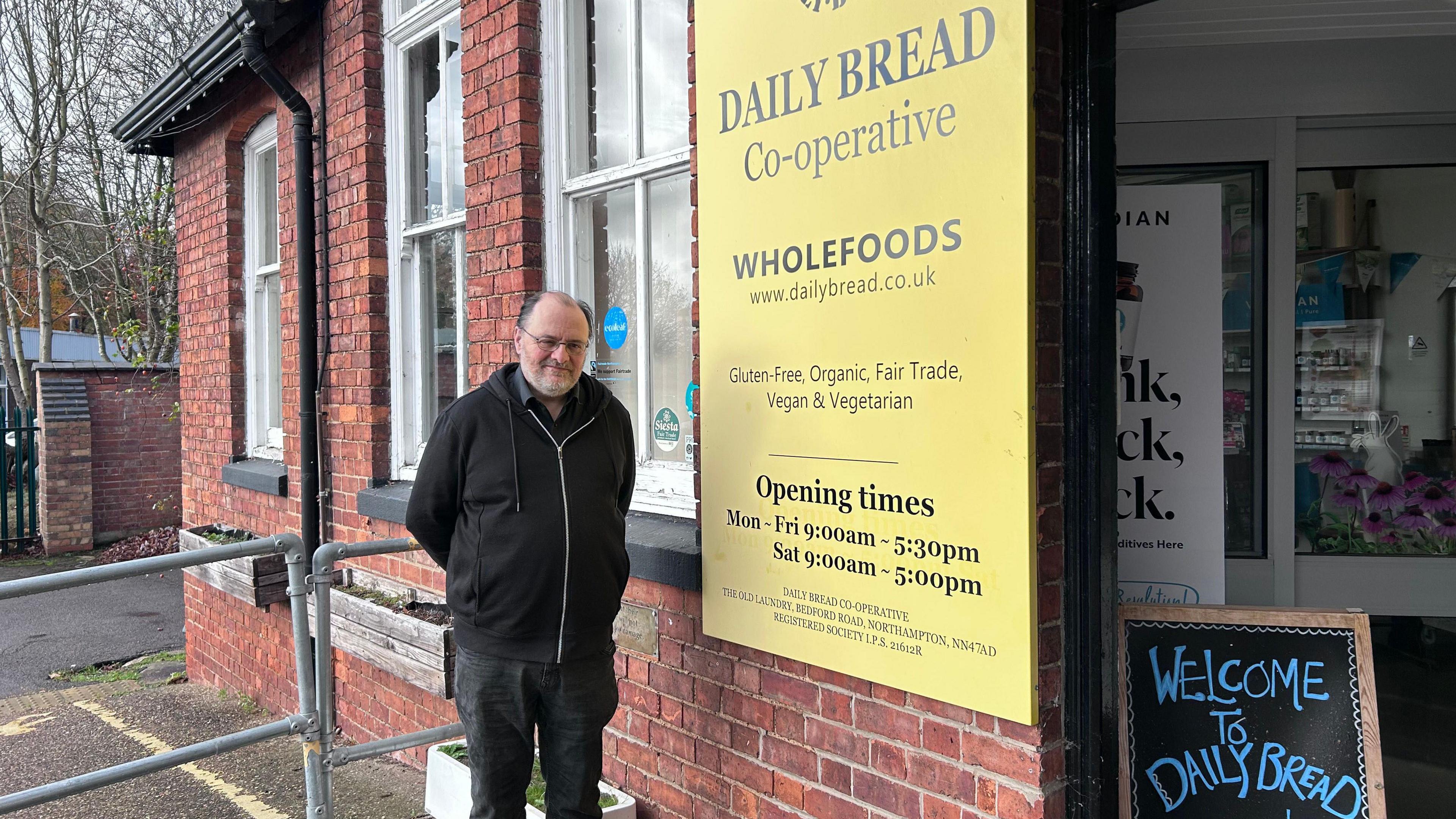 John Clarke, who is a member of the Daily Bread co-operative health food shop. He stands outside his shop, which is to the front of the red brick Victorian Workbridge building. To his left there is a large yellow shop front sign, with the logo which is bread shaped. It advertises its opening times. There is also a chalk board saying Welcome to Daily Bread.