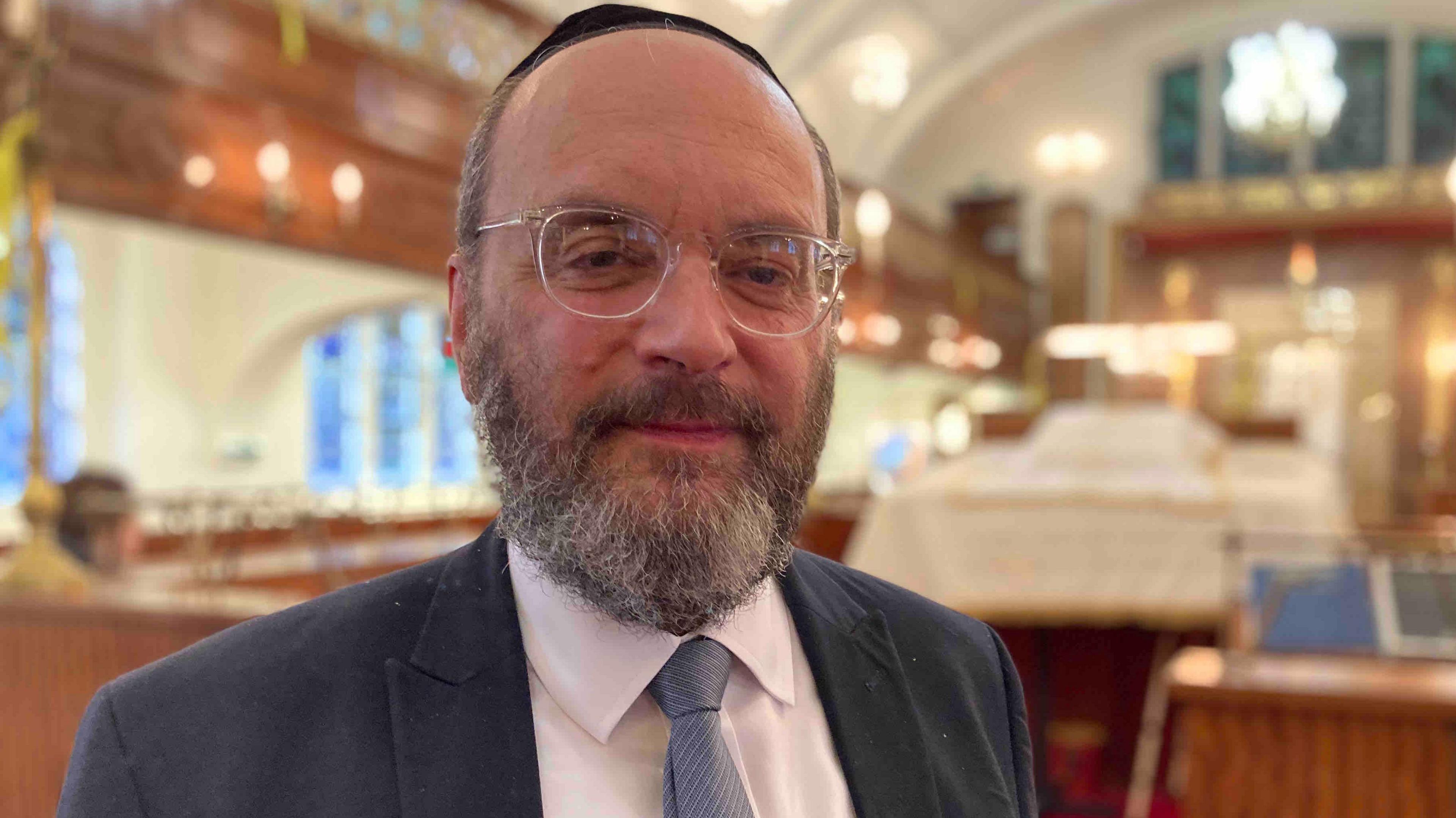 Rabbi Alan Lewis is looking at the camera inside a synagogue. He is wearing a suit, cap and glasses. His tie is blue. 