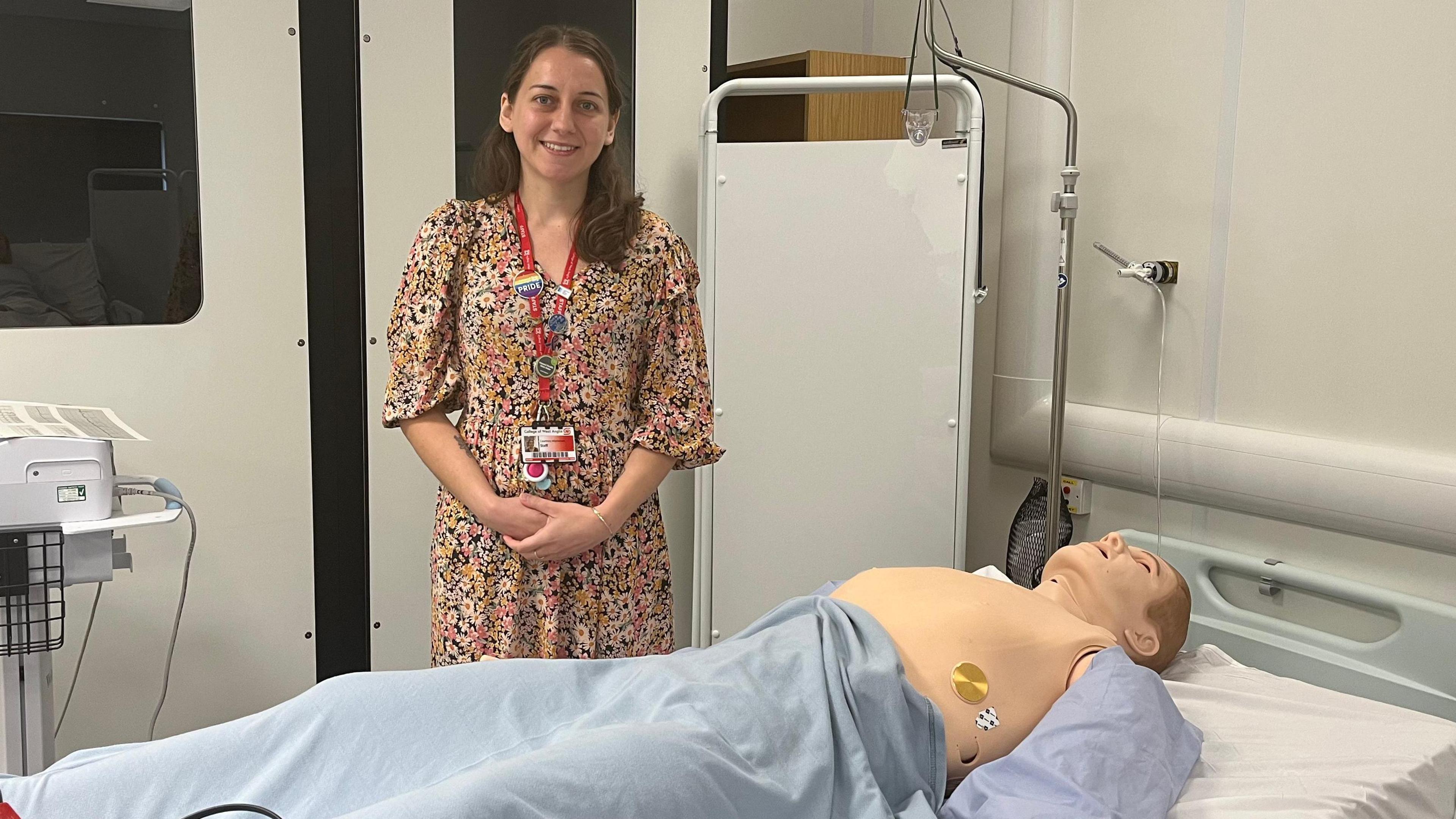 Courtney Masterson is standing in front of a hospital bed with a dummy in front of her wearing blue scrubs. Courtney is wearing a flower patterned dress and has brown hair. Her hands are folded in front of her.