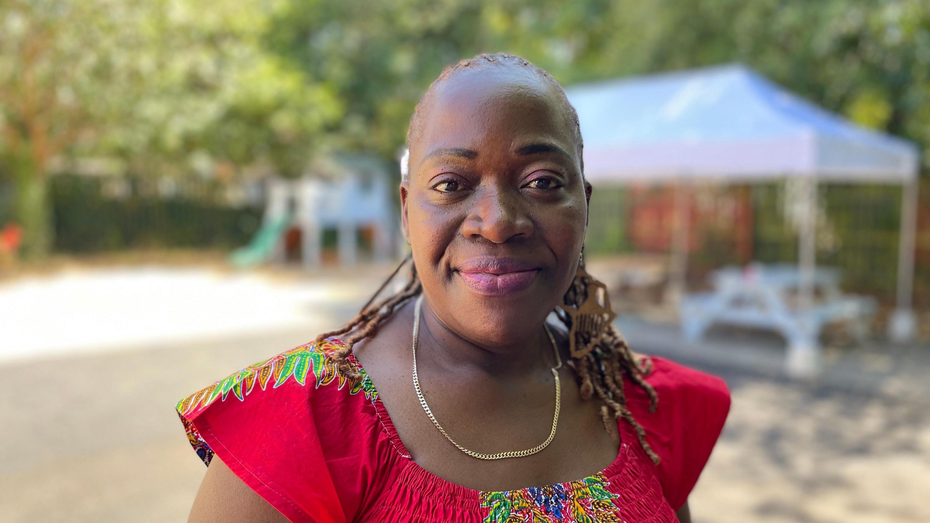 Bernadetta Omondi is pictured from her shoulders up wearing a red top with blue, yellow and green patterns on the centre and shoulders. She wears a gold necklace and is smiling at the camera. The background is a blurred so the camera is just focused on her.