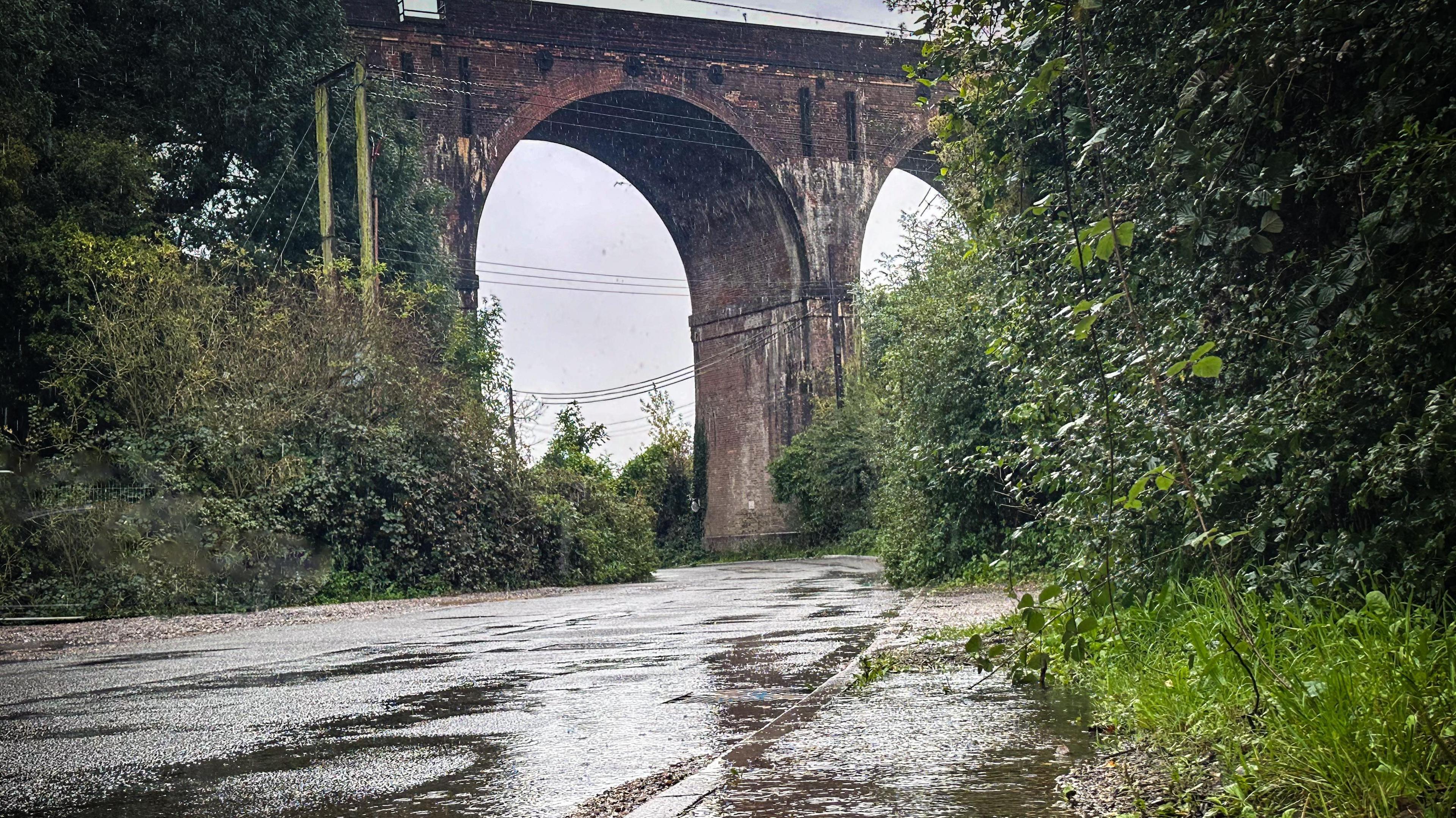 A brick arch viaduct is photographed over a road. It is raining and there are large puddles on the road. There is a green hedge on either side and the sky above is a dark grey.