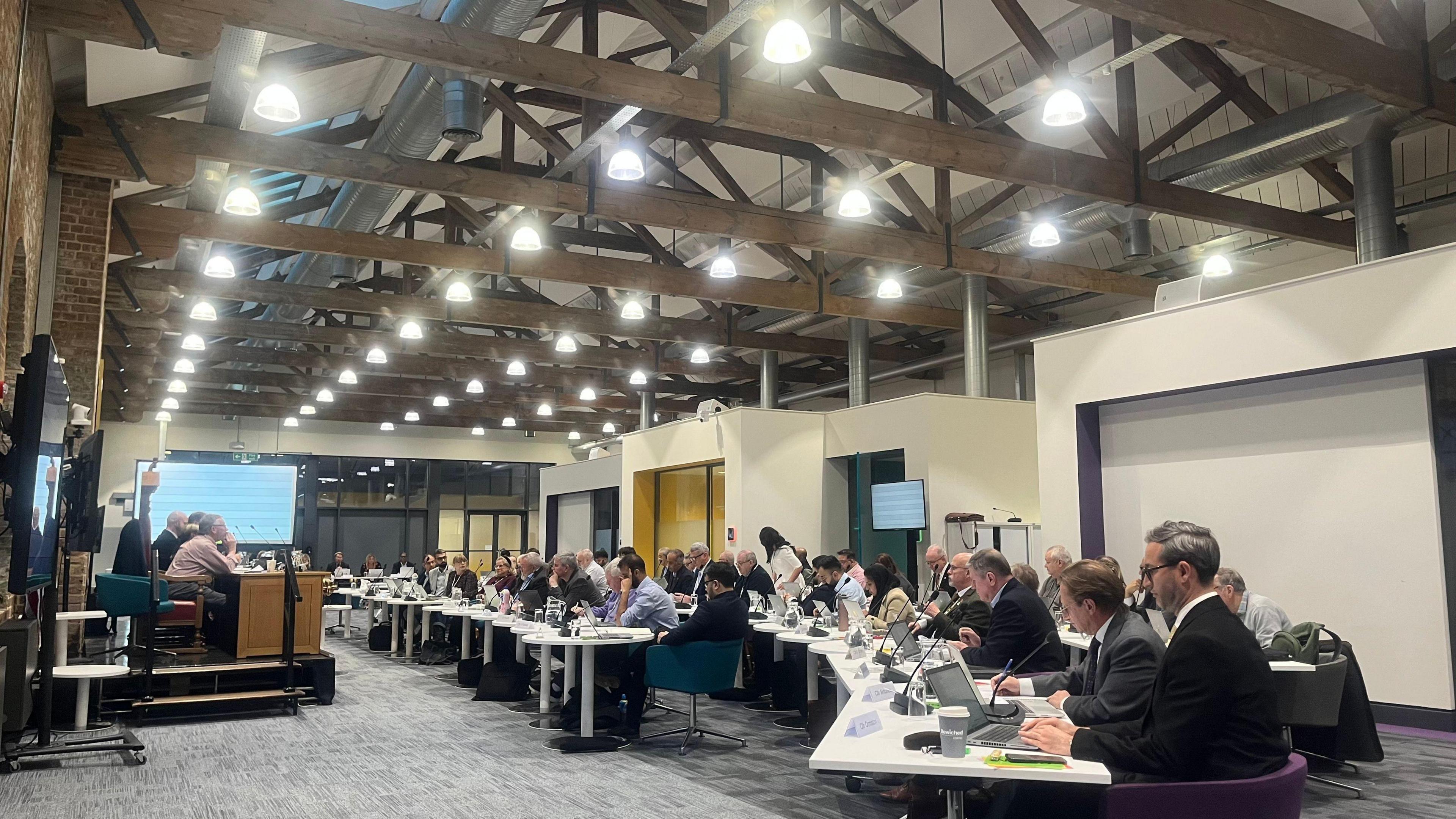 A council meeting room- with councillors sitting in rows on white tables.