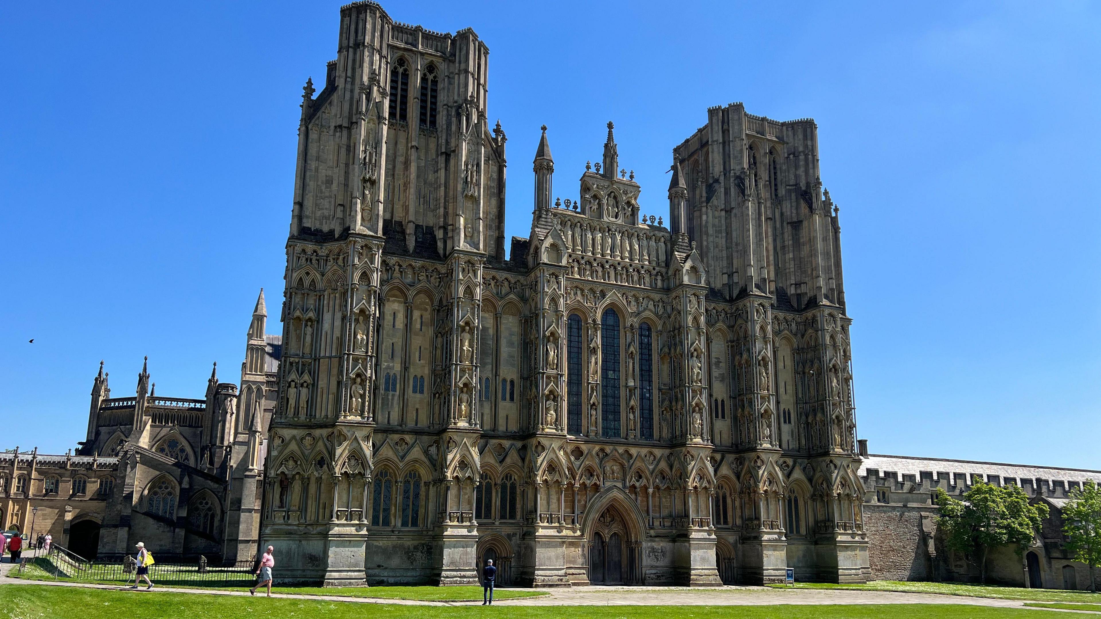 A picture of the outside of Wells Cathedral on a sunny day.  People are walking outside.