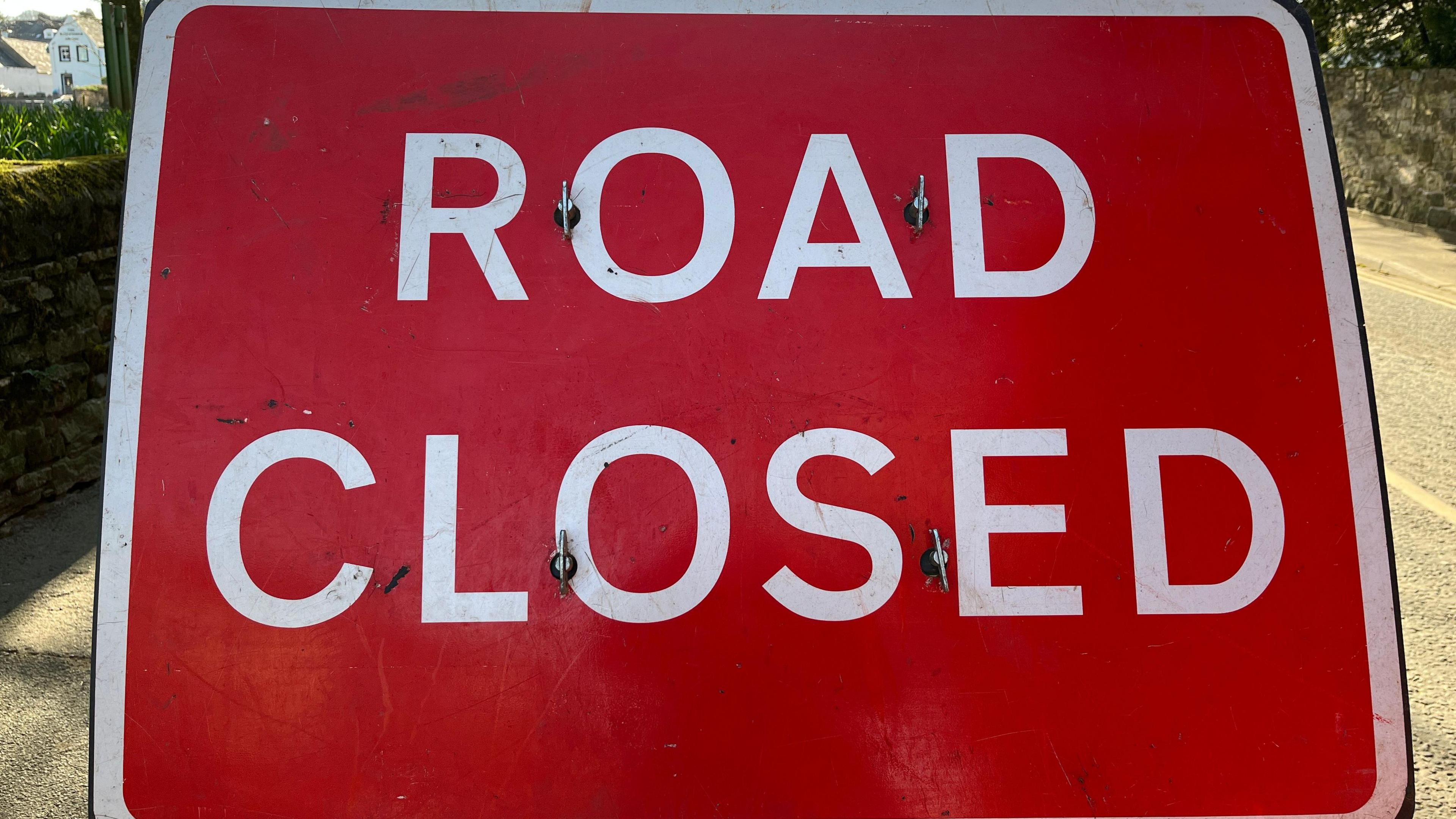 A Road Closed sign with white writing and a red background, a white border, zoomed in so you can only see parts of the road at the side of the sign.