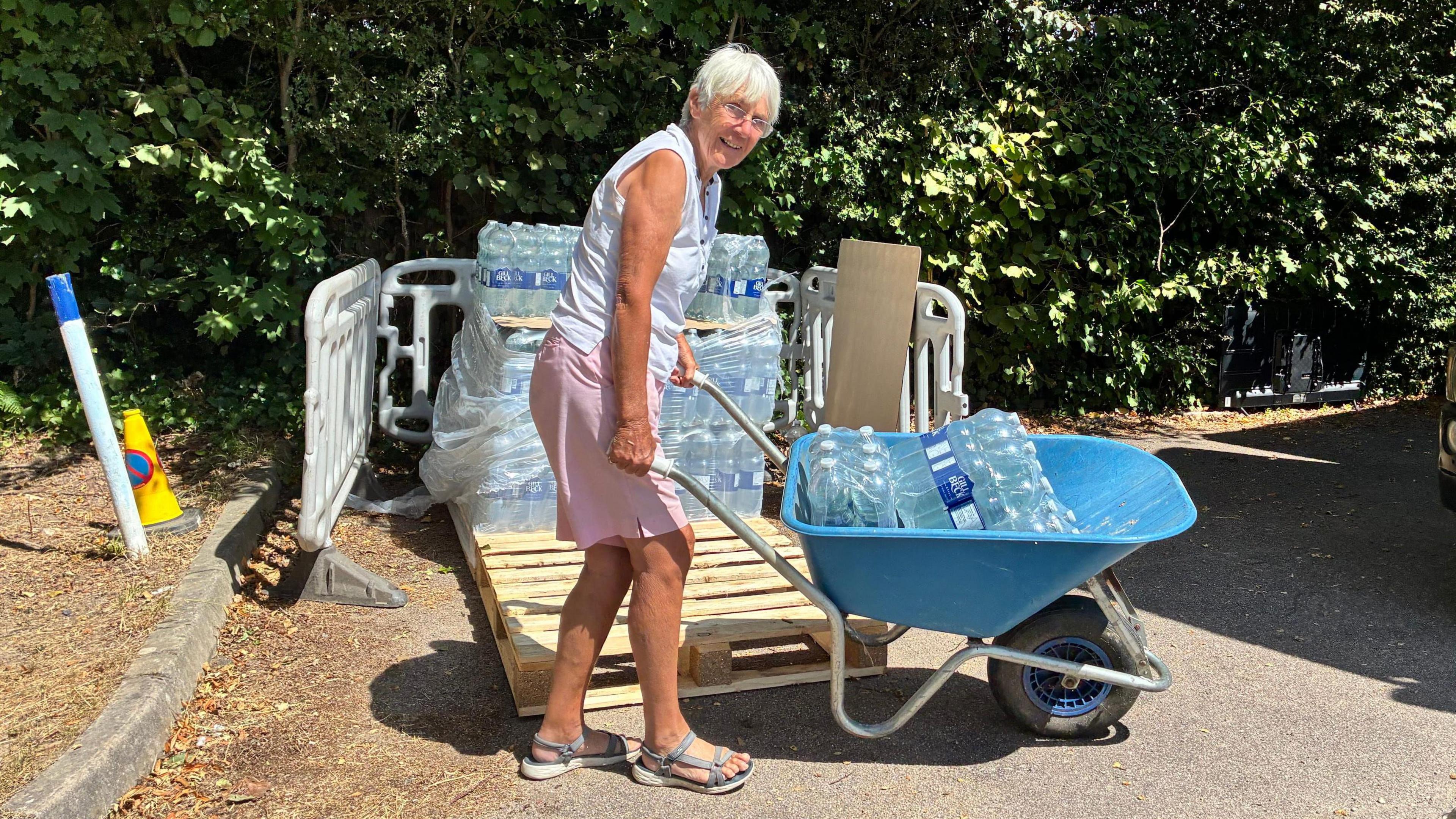 An elderly woman carrying crates of water in a wheelbarrow