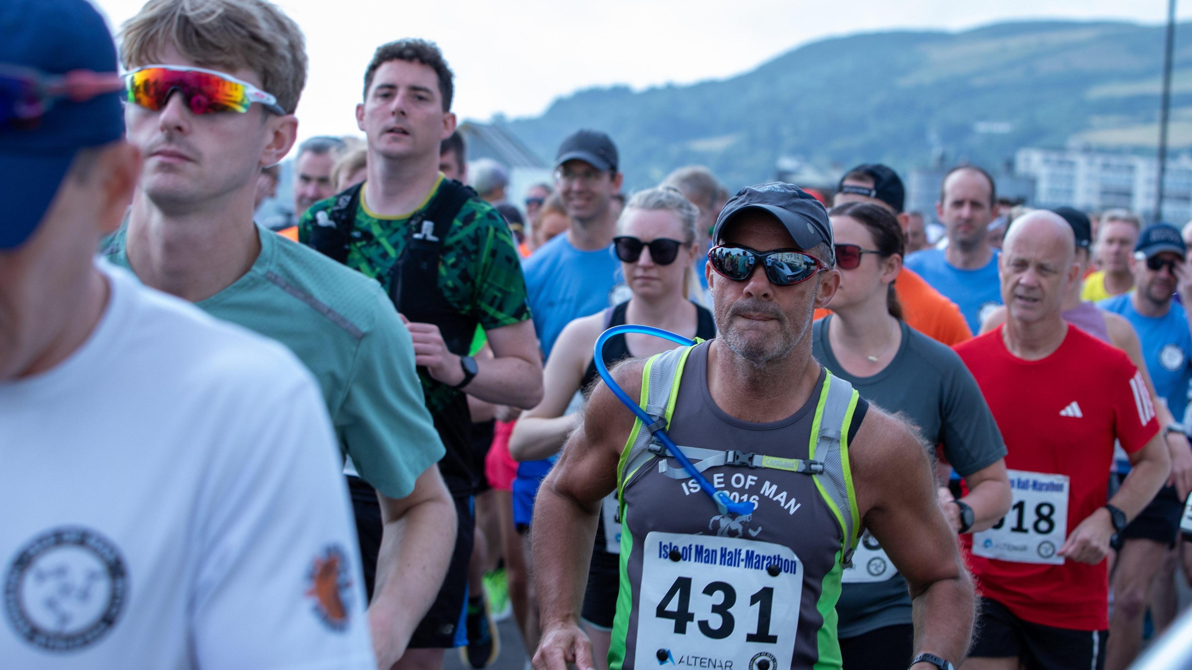 A group of people running with numbers on their torsos. One man wearing a cap and blue sunglasses runs past, you can see the hill in the background in Ramsey.