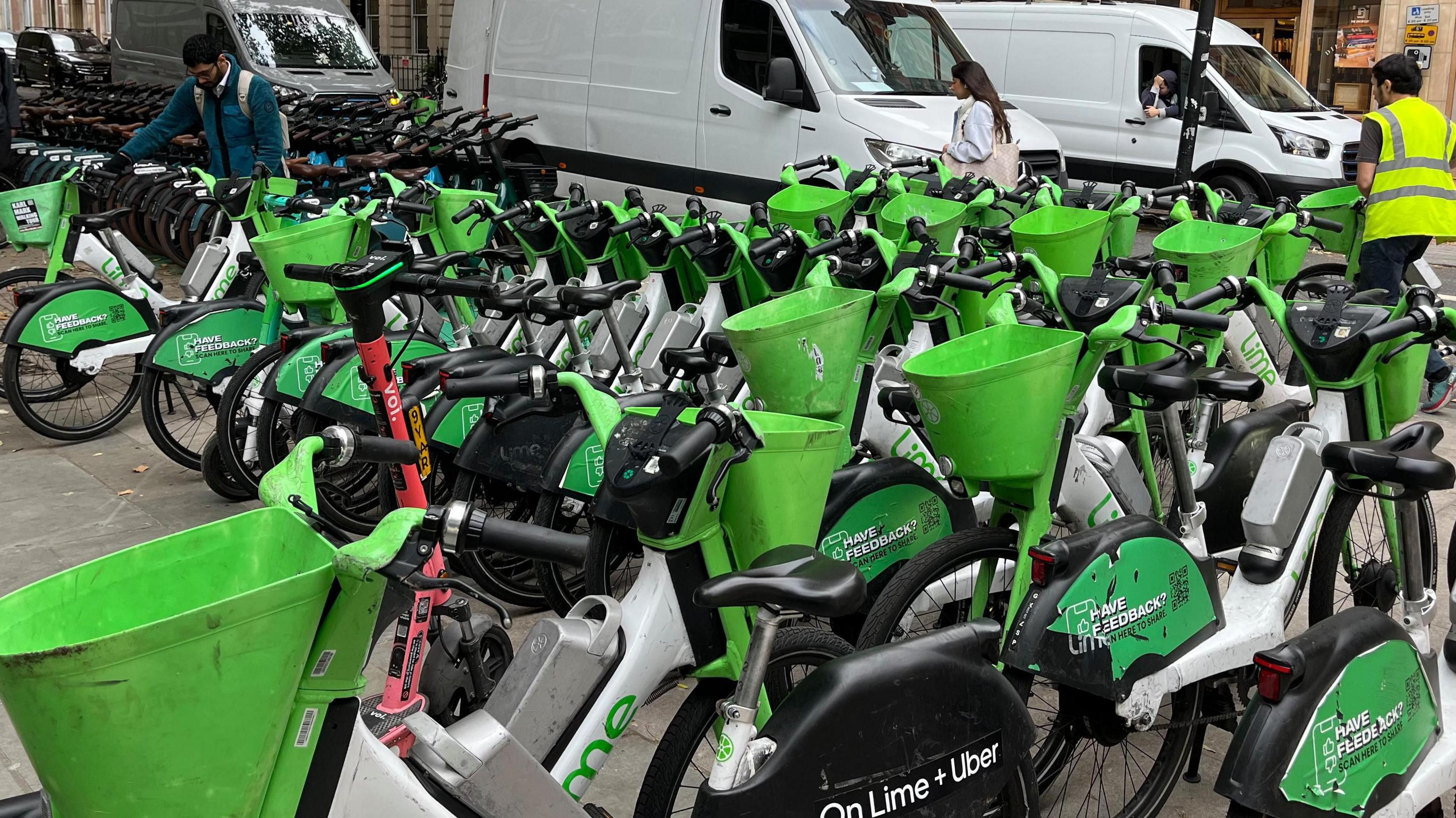 A group of Lime bikes are on the pavement and a man in a hi viz is moving one into the back of a parked white van