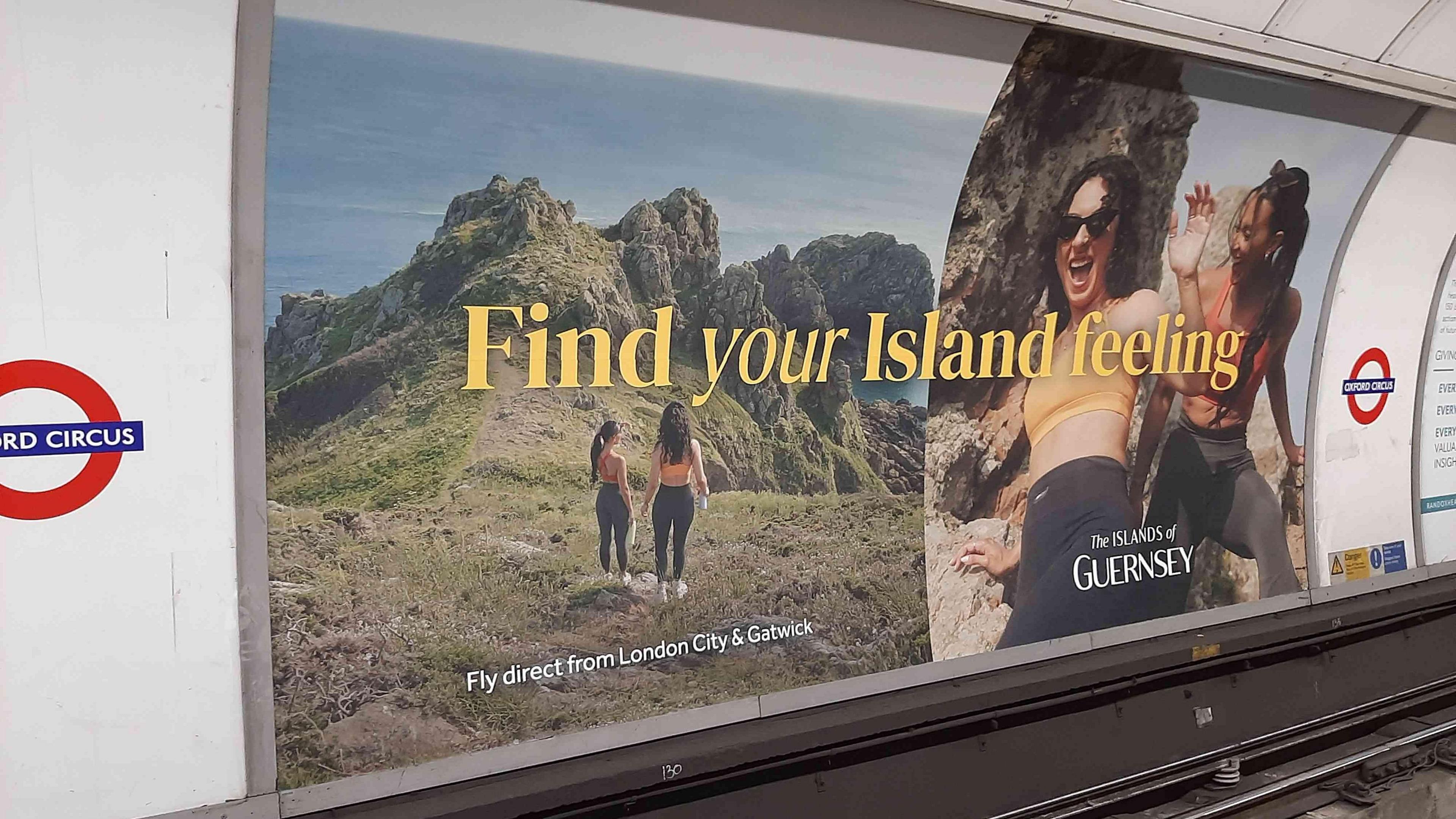 A London Underground advertising billboard at Oxford Circus station. Pictures of two women in coastal areas of Guernsey by some cliffs. Text reads: Find your island feeling. Fly direct from London City and Gatwick. The Islands of Guernsey.