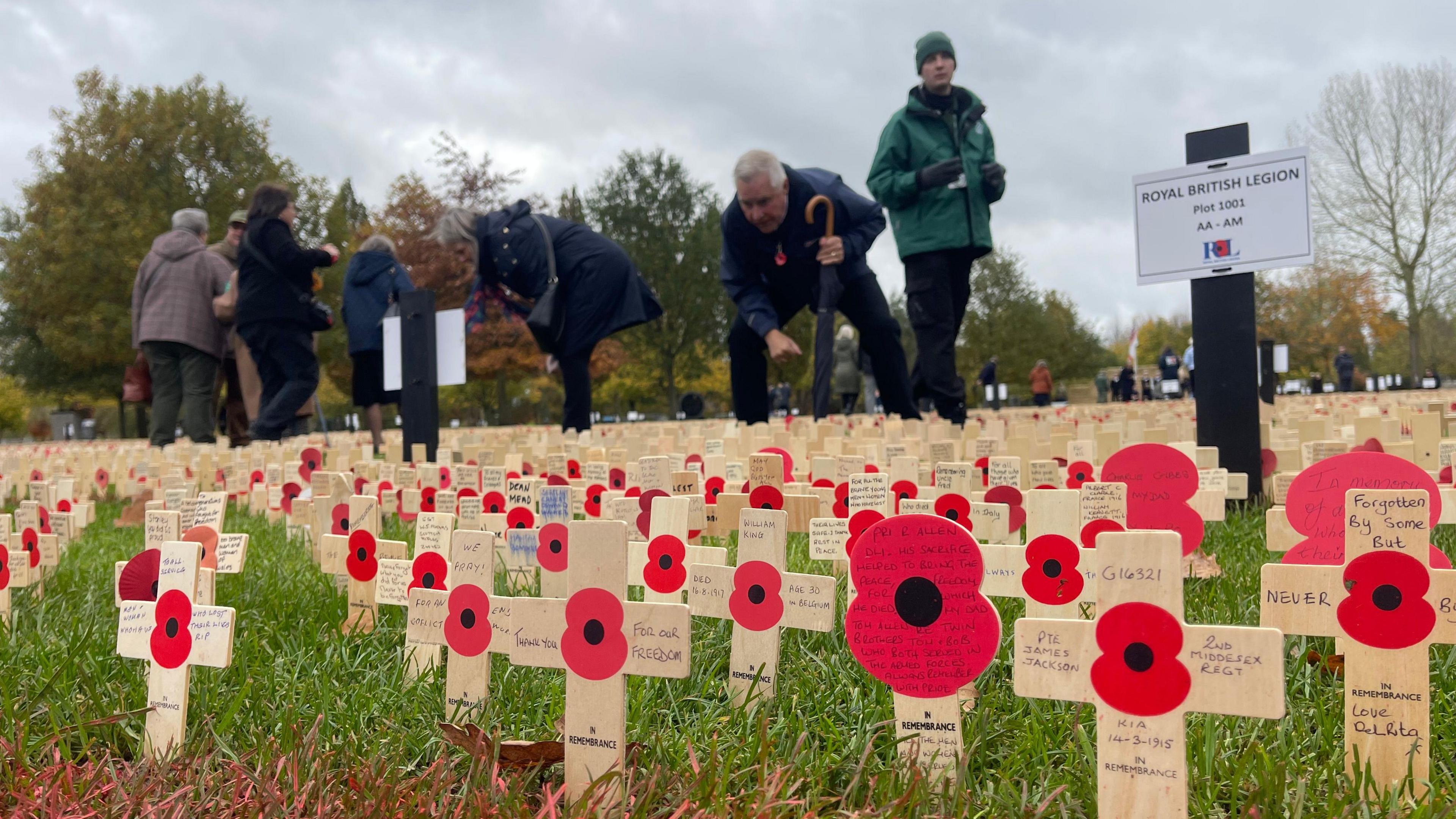 Hundreds of wooden crosses which each bear the image of a red poppy and a handwritten note. They are placed in the grass, while people in the distance are reading some of the messages.