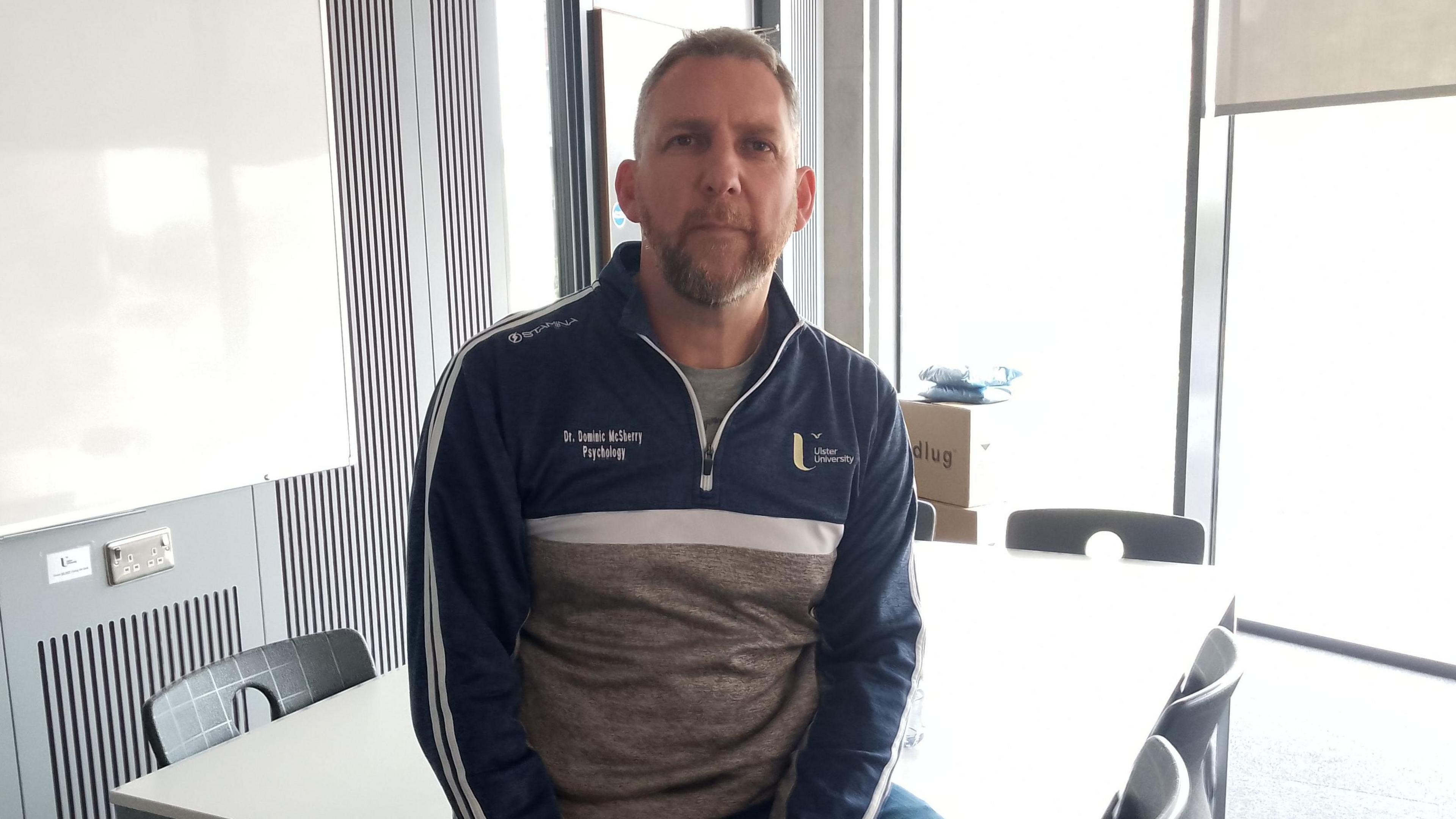 A man with light hair sits on a classroom table and looks at the camera. He is wearing a navy and grey half zip jumper with the Ulster University logo embellished on the front