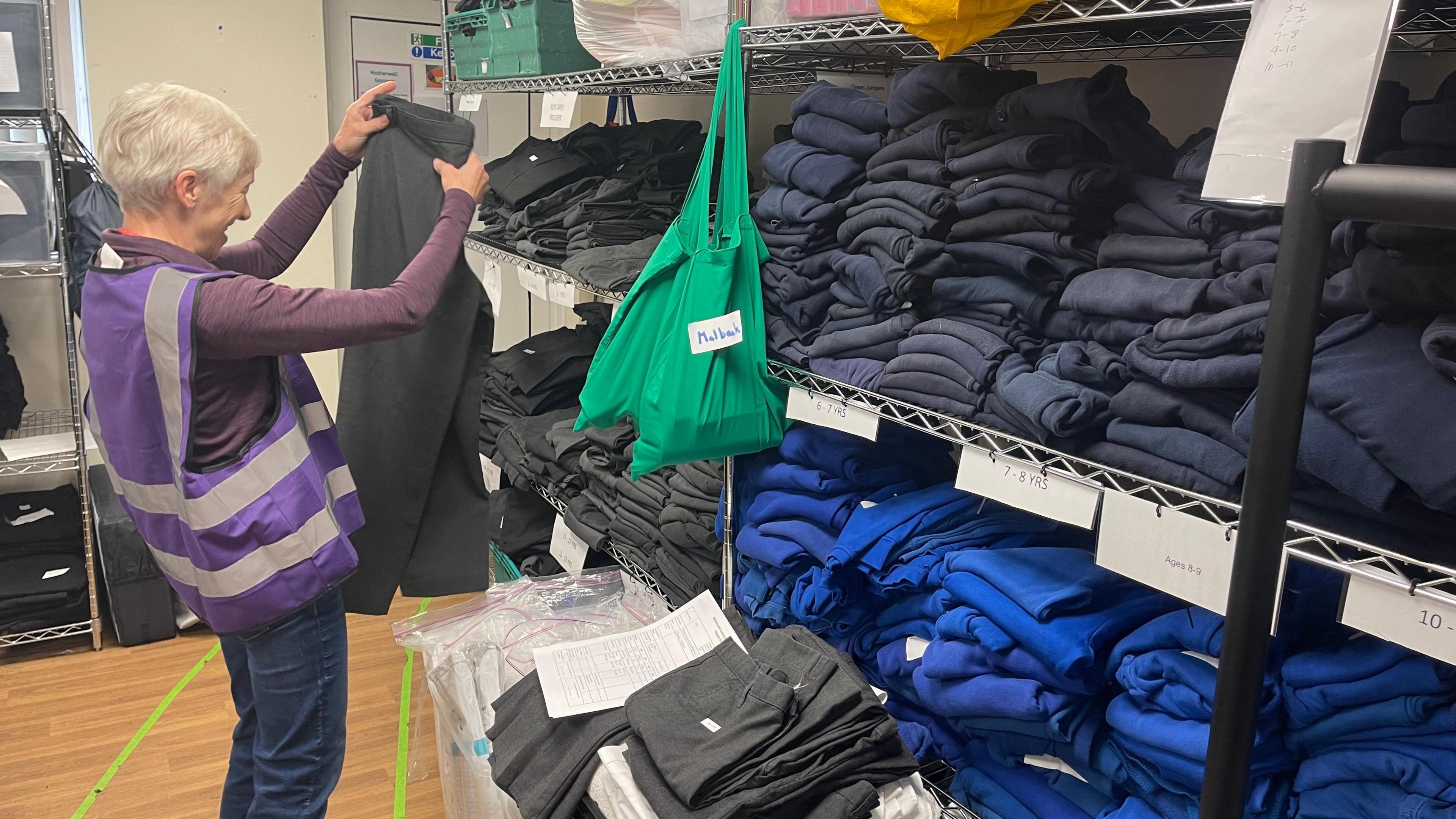 An elderly lady in a purple hi-vis folds a pair of great school trousers. She stands beside shelves packed with other school uniform items.