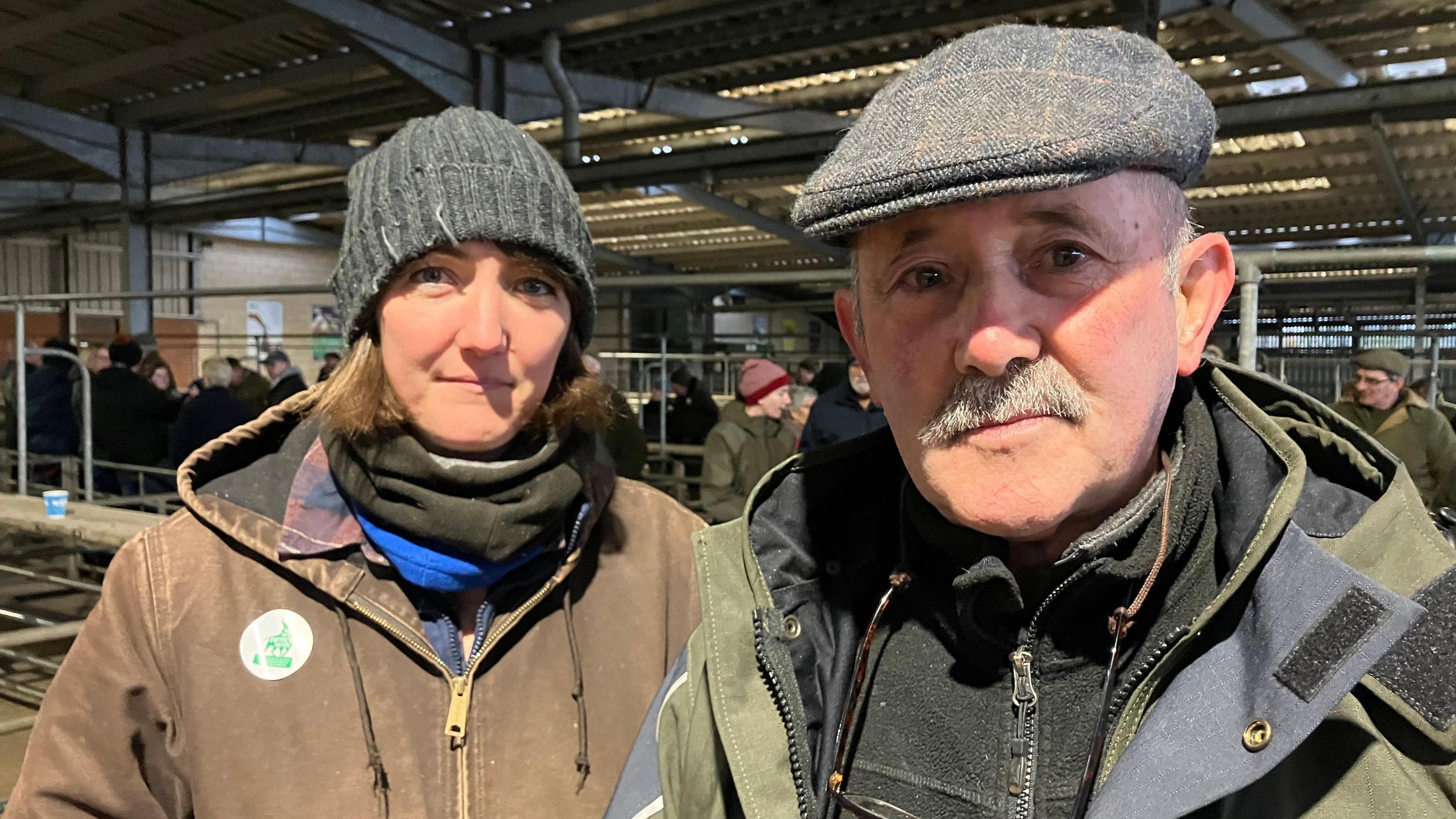 A woman and man stand in an agricultural auction house. The woman, who is on the left, is wearing a wooly hat and a brown coat. The man, who is on the right, is wearing a grey flatcap and a green jacket with a black fleece underneath.