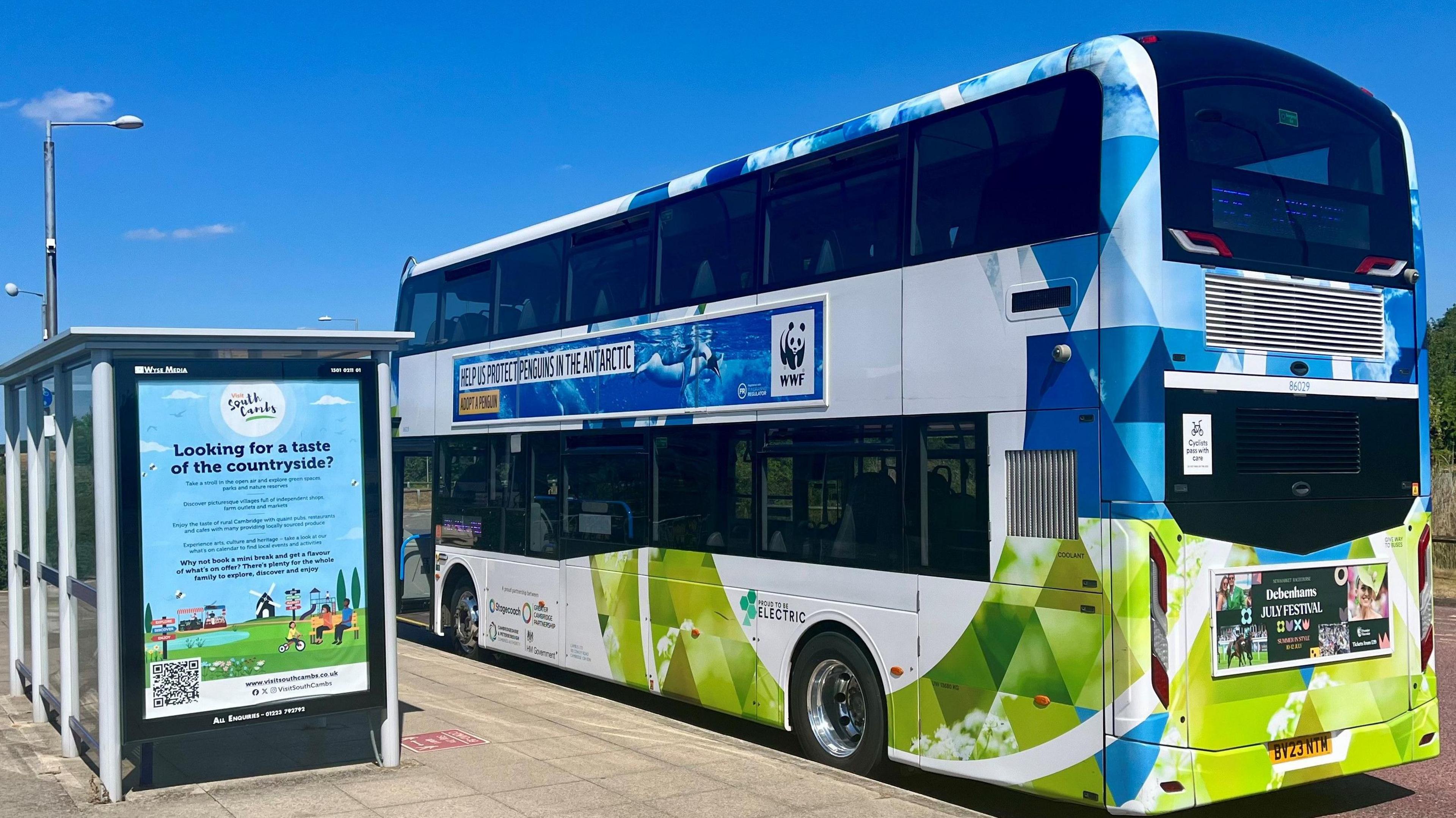 A colourful bus is parked at a bus stop. It is a double decker and it is white, blue and green. It has a WWF advert on the side with the charity's black and white panda logo. The bus also has a sign indicating it is an electric vehicle