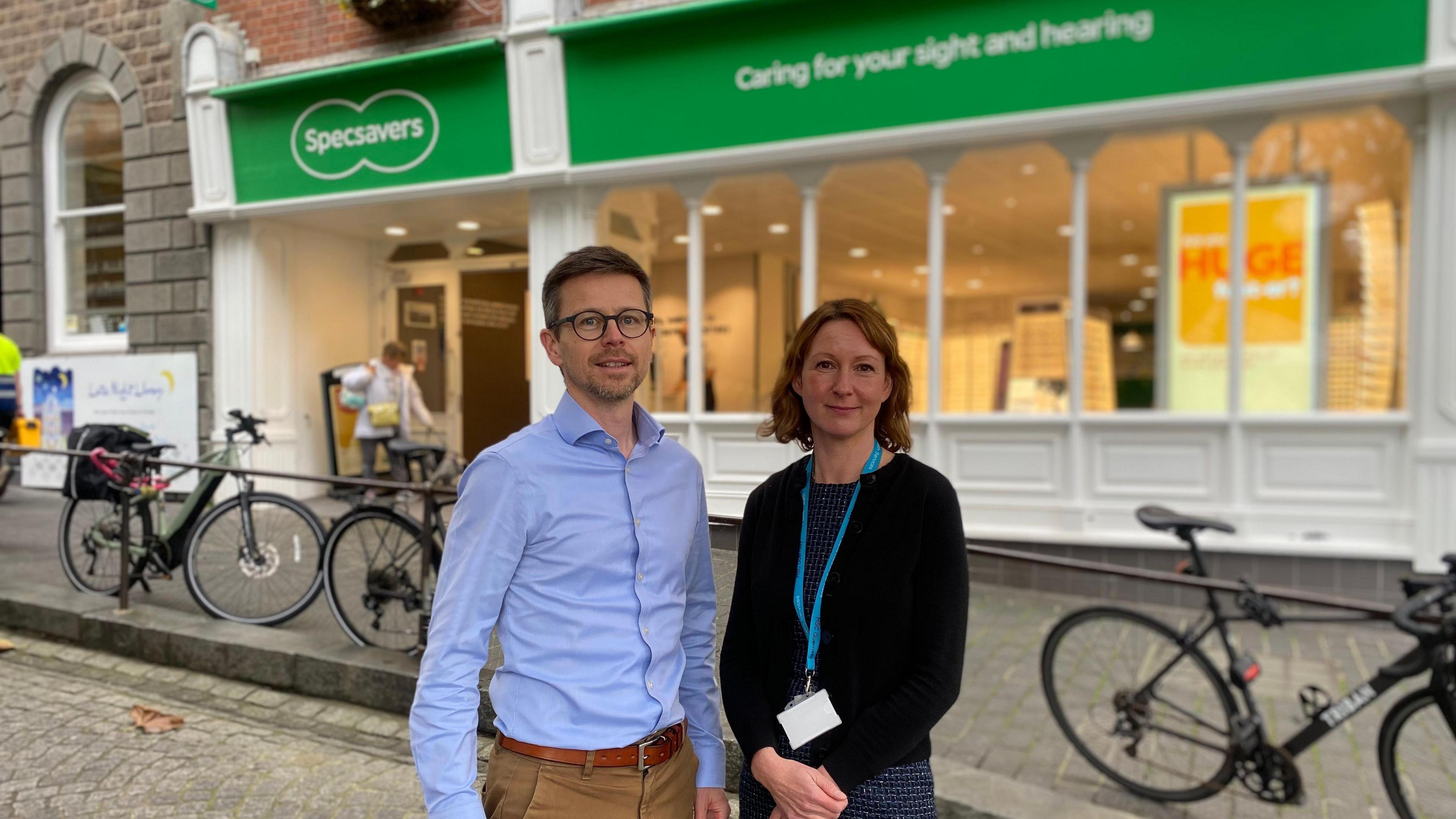 A man with brown hair in a blue shirt with camel brown trousers and a woman with brown hair and a blue dress stand outside of Specsavers opticians in Guernsey's Market Square