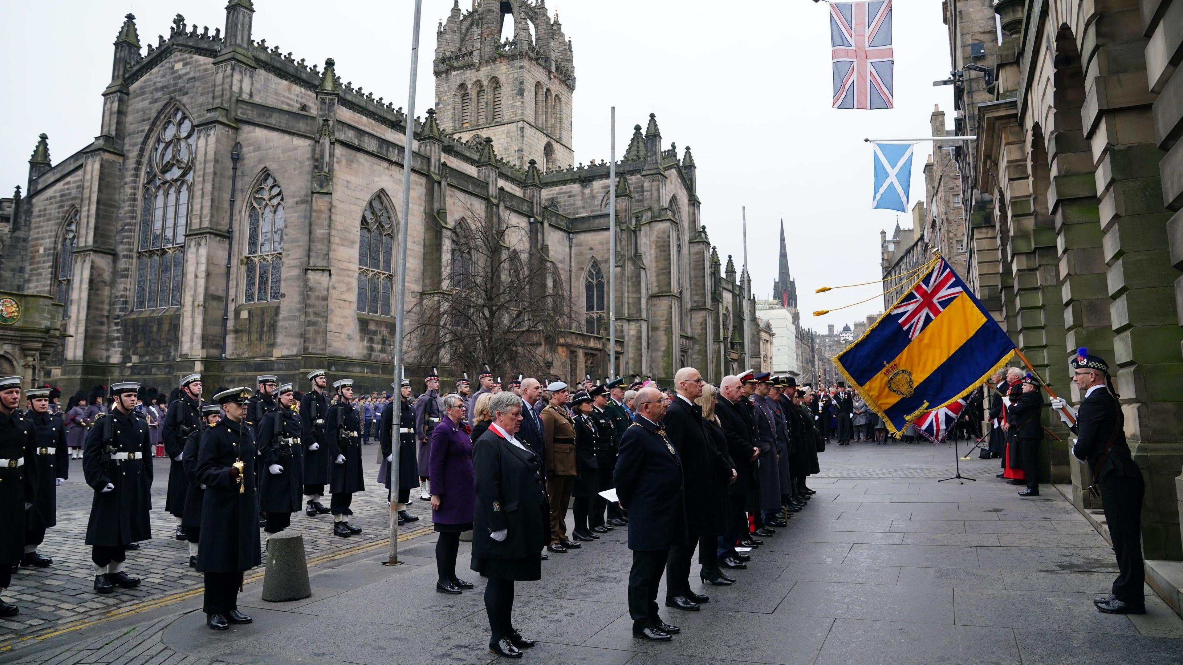 People stand in rows, many in military uniform, all facing Edinburgh's City Chambers, which has a UK and Scottish flag hanging from it. St Giles' Cathedral is behind them.