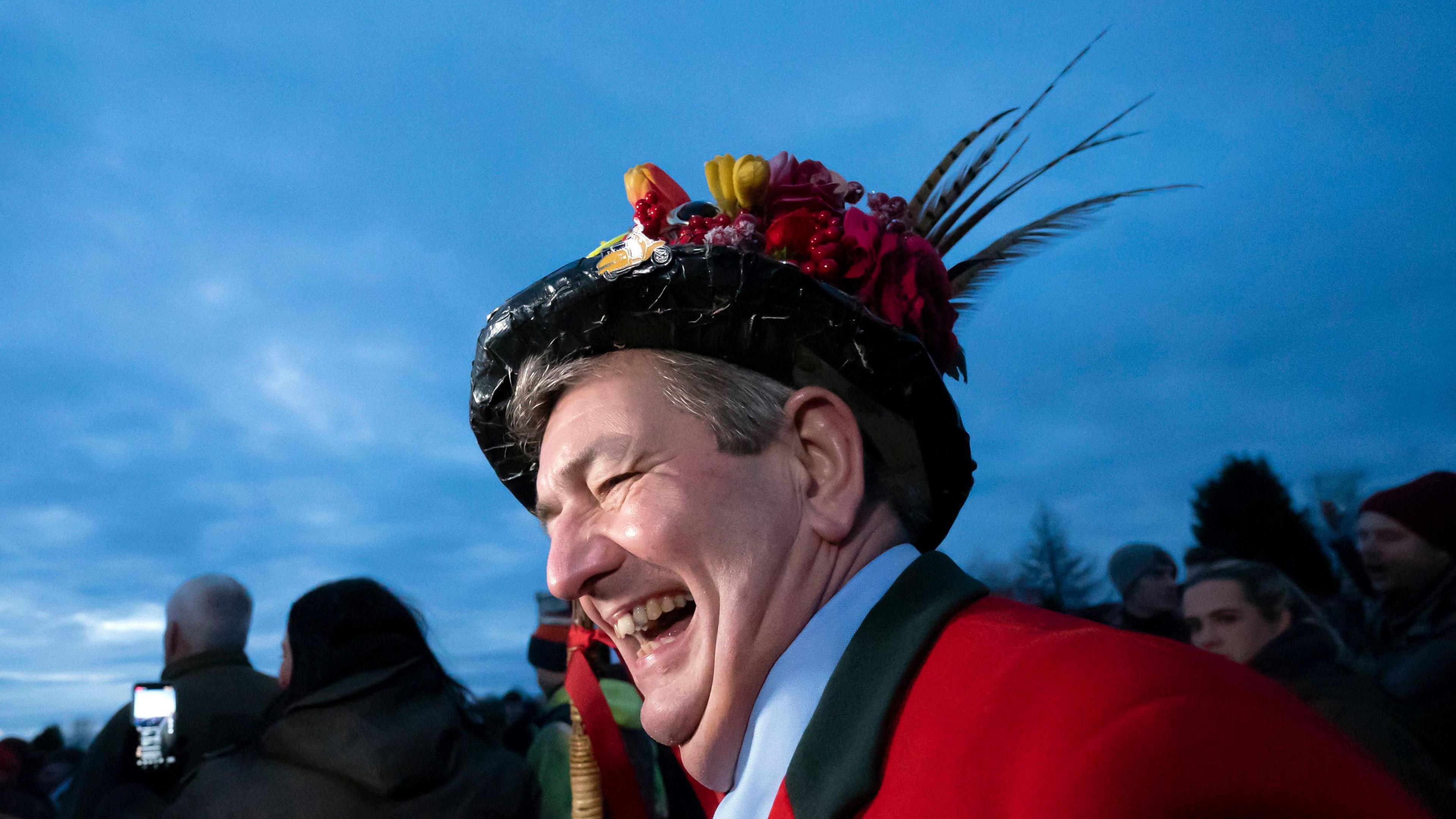 A man laughing to the side wearing a black top hat with colourful flowers and feathers on it and a red blazer. It is night-time and you can see other people stood in the background wearing coats.