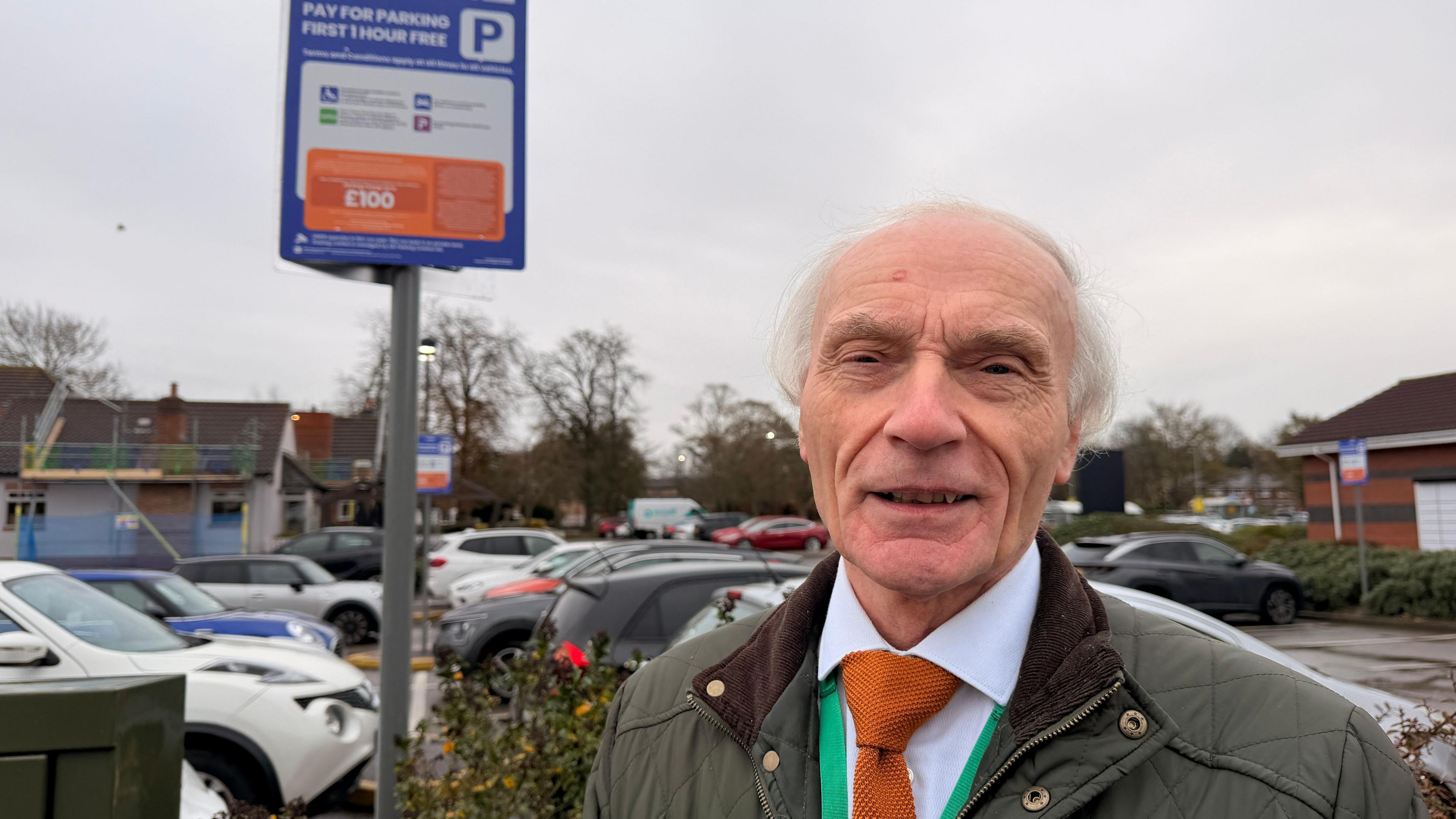 A man with white hair in a suit, standing in a car park next to a large sign where there is small print with instructions on how to park 