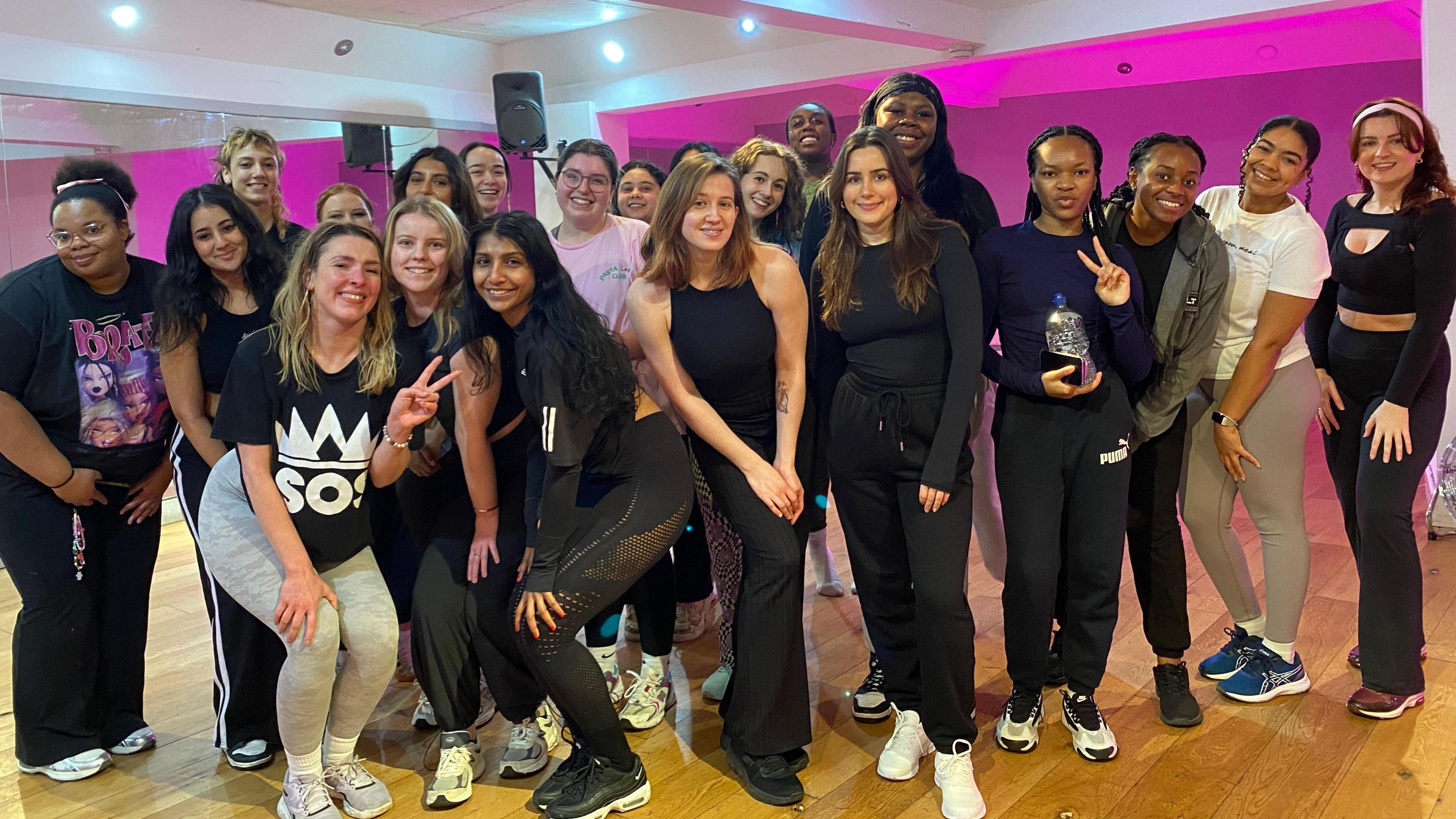 A group of around 20 women are standing together and smiling. They are wearing leggings and tops with some holding water bottles. Bright pink lights shine in the background of a dance studio