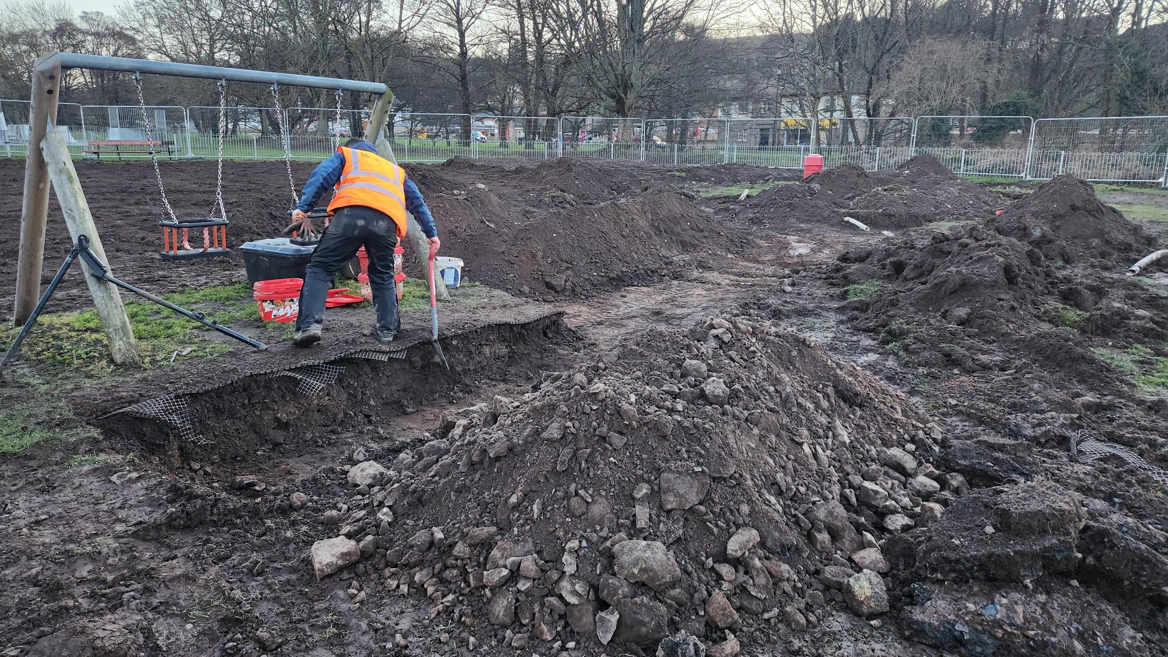 A man digs in front of a pair of swings. Around him are large mountains of earth in a play park which has been almost completely dug up 