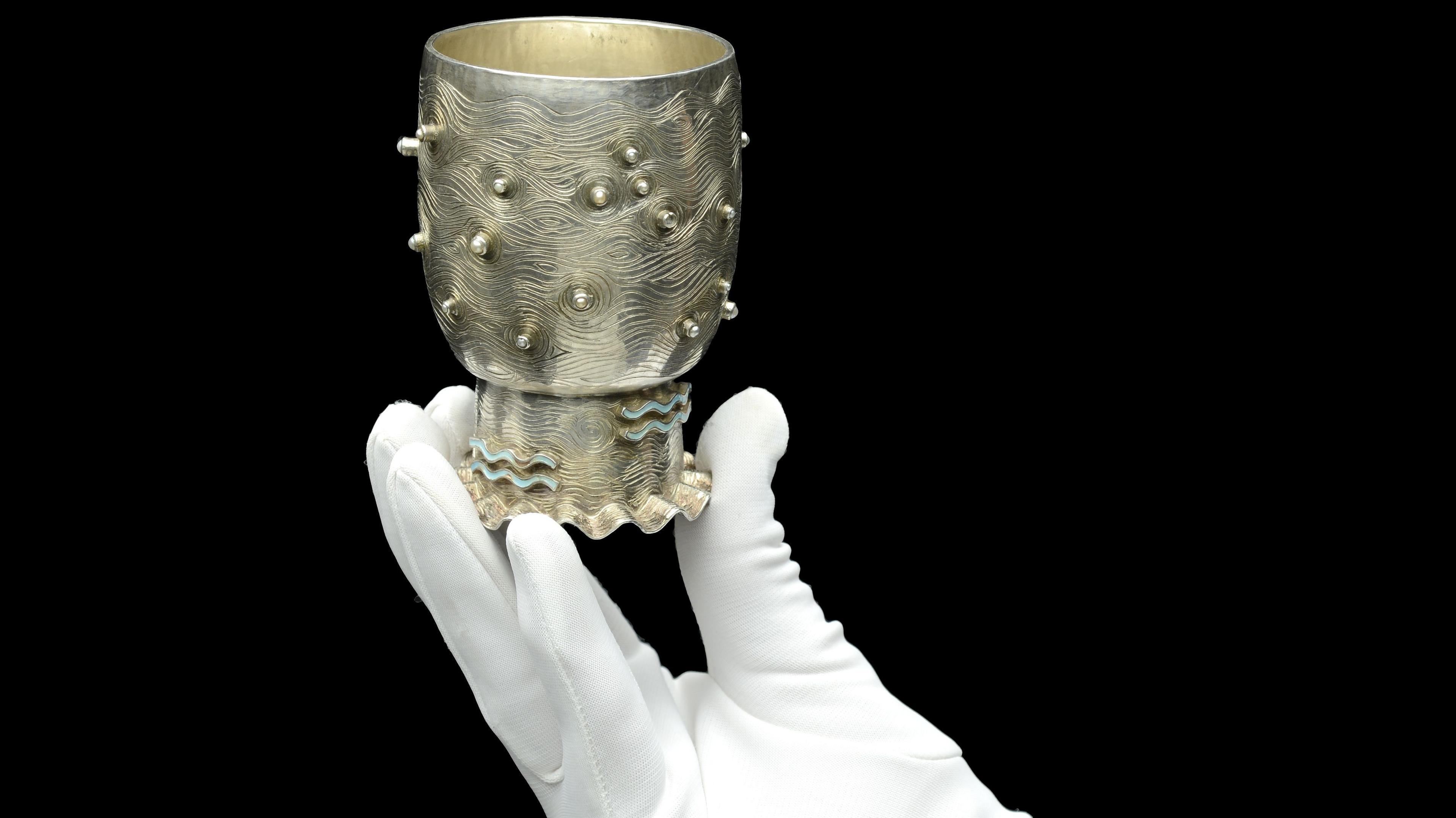 A small silver goblet being held by a white handed glove on a black background 