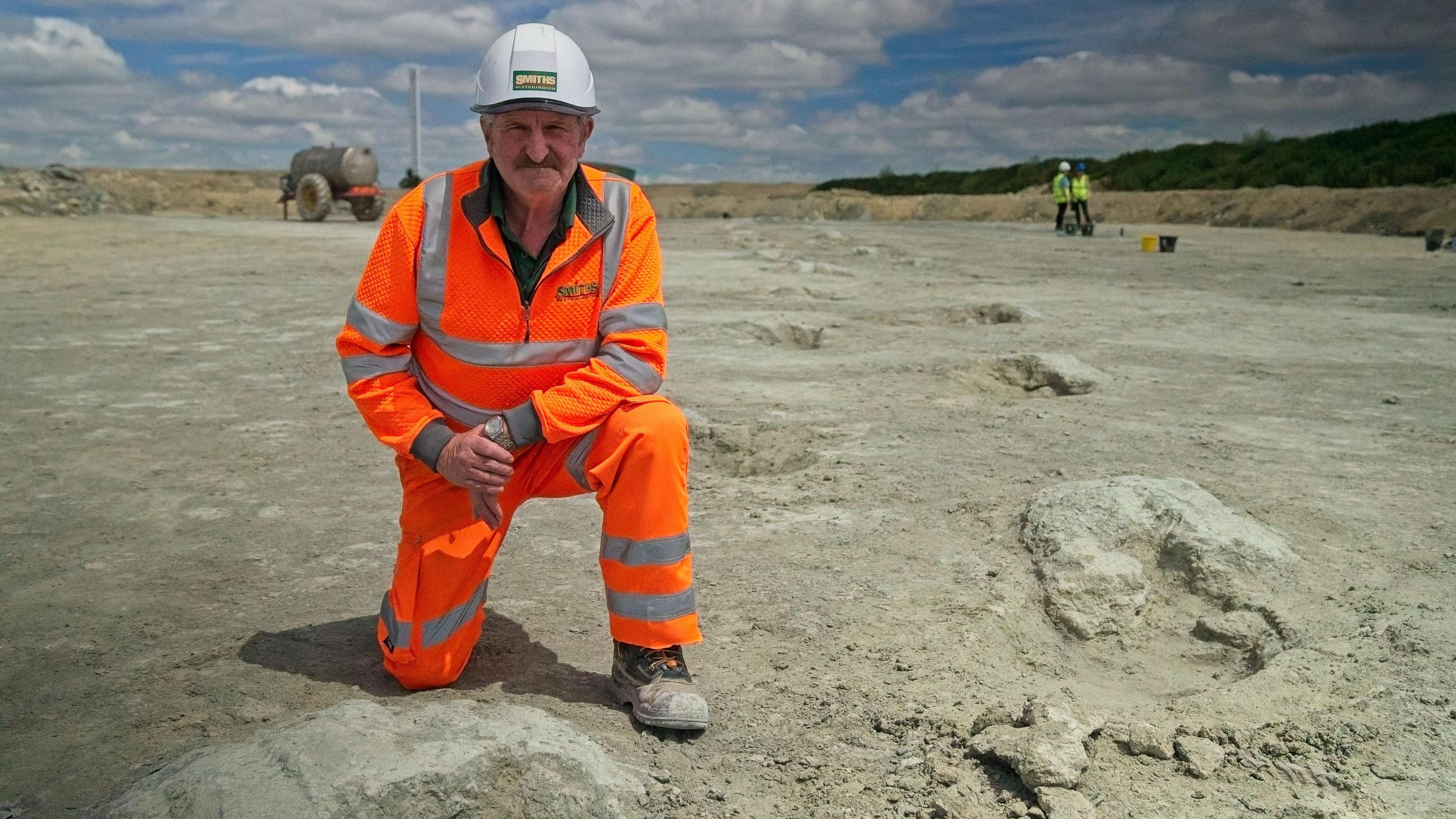 Gary Johnson a man looking to be in his sixties with a determined state and a grey moustache, dressed in an orange jumpsuit and sand covered boots with a white helmet, kneels with one knee up, one knee on the ground next to the dinosaur footprints he found. They are large craters of indistinct shape in this picture, which trail off into the distance in the whitish-grey sand of a quarry. In the distance behind him to the right two people in yellow hi-visibility waistcoats and hard hats stand with buckets beside them on the ground.