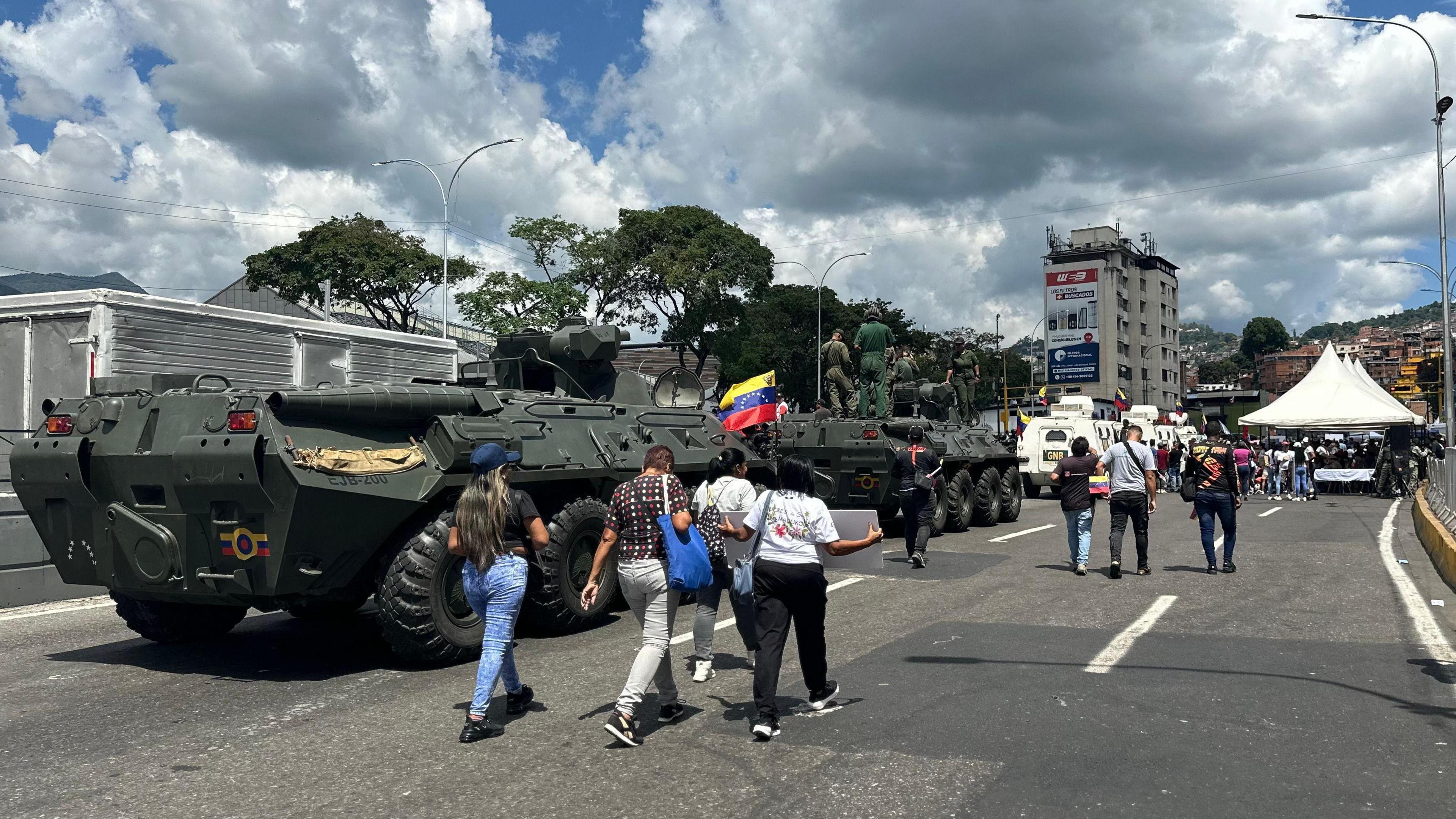 People walk past armoured vehicles in a street in the capital, Caracas. In the background, tents can be seen. 
