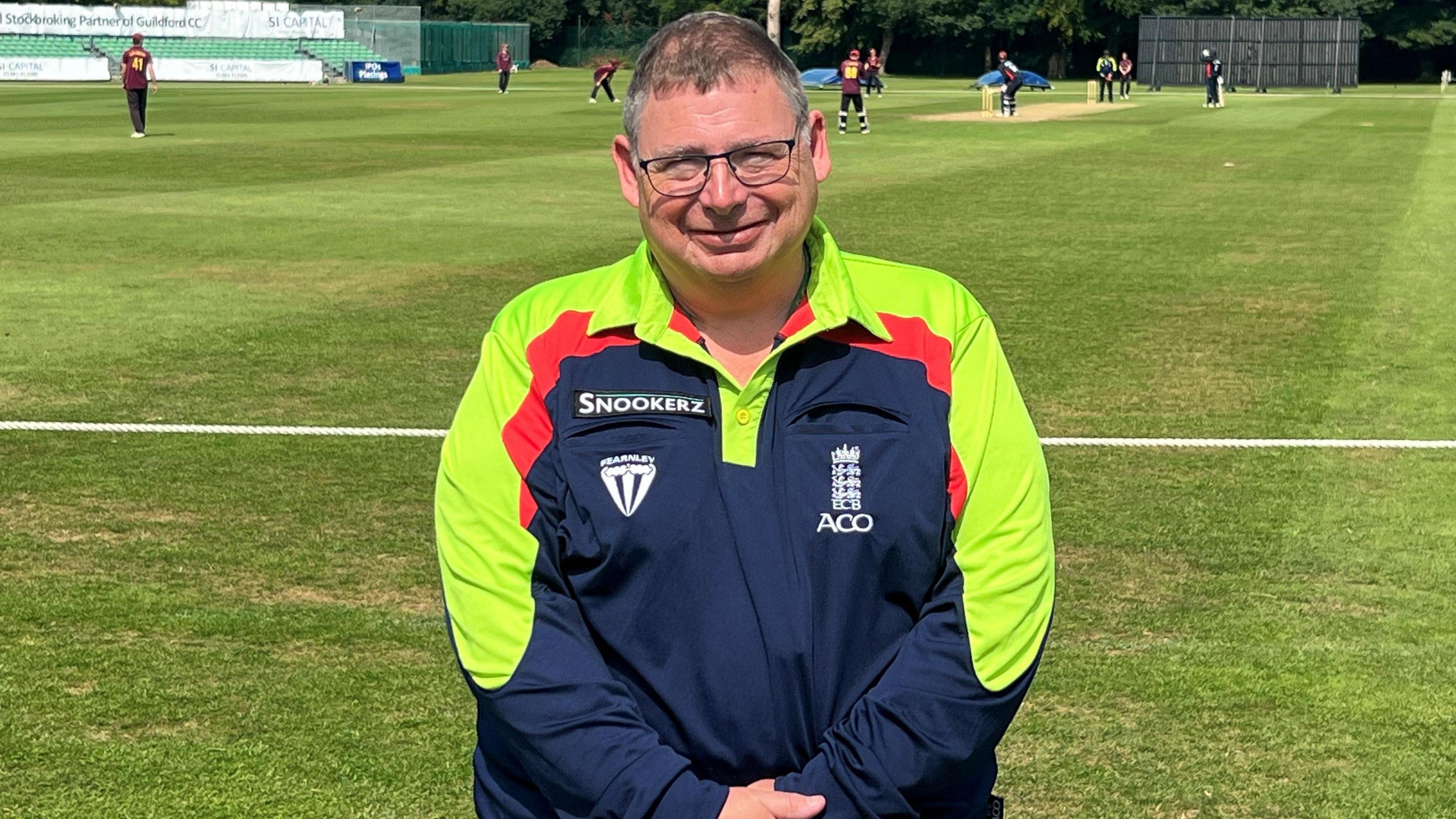 Terry wearing glasses in a navy England Cricket Board shirt with bright green sleeves in front of two teams playing cricket