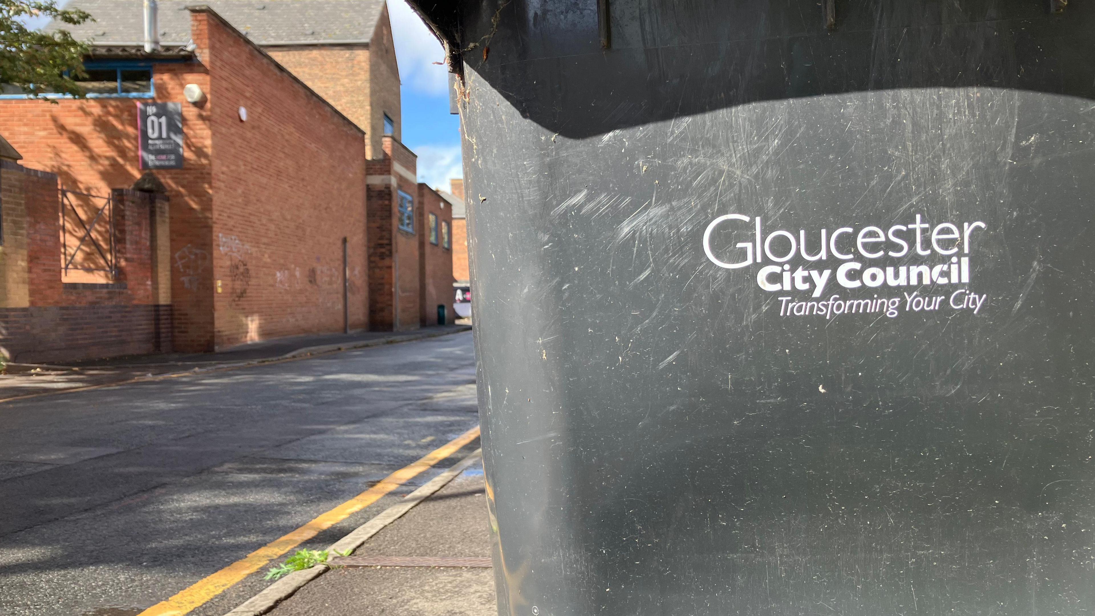 The image captures a typical urban street scene in Gloucester. Dominating the foreground is a large black wheelie bin bearing the label “Gloucester City Council Transforming Your City”. Behind the bin, brick buildings line the street. The overall setting is residential
