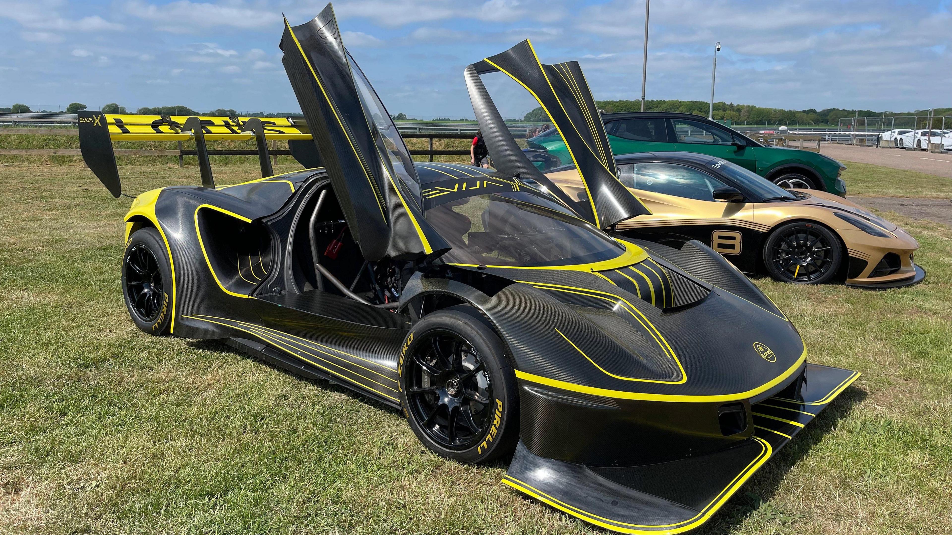 A black and yellow sports car is parked on grass with its doors open alongside other cars