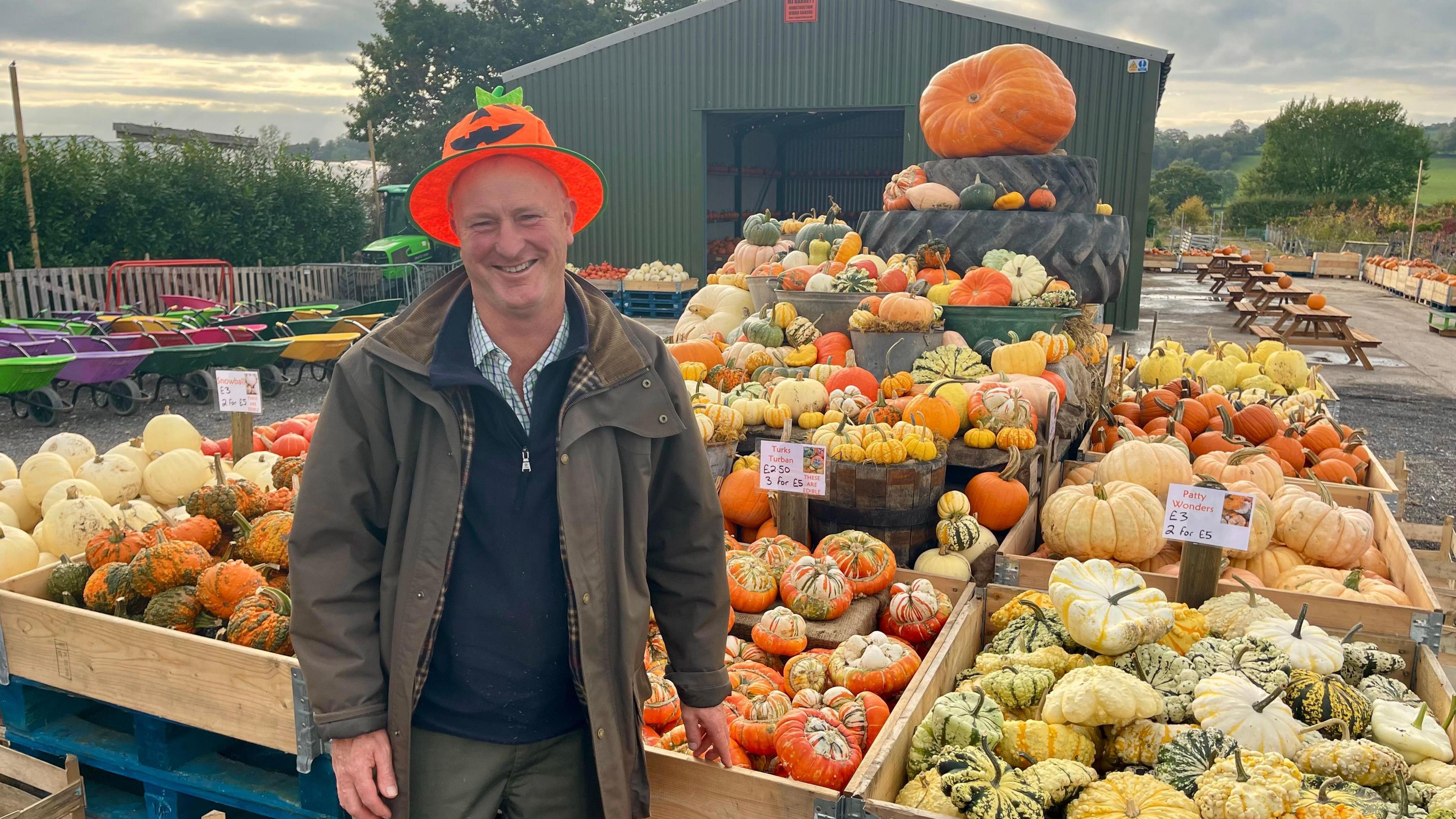 A man in a pumpkin hat is standing in front of over a dozen crates of pumpkins of different varieties.