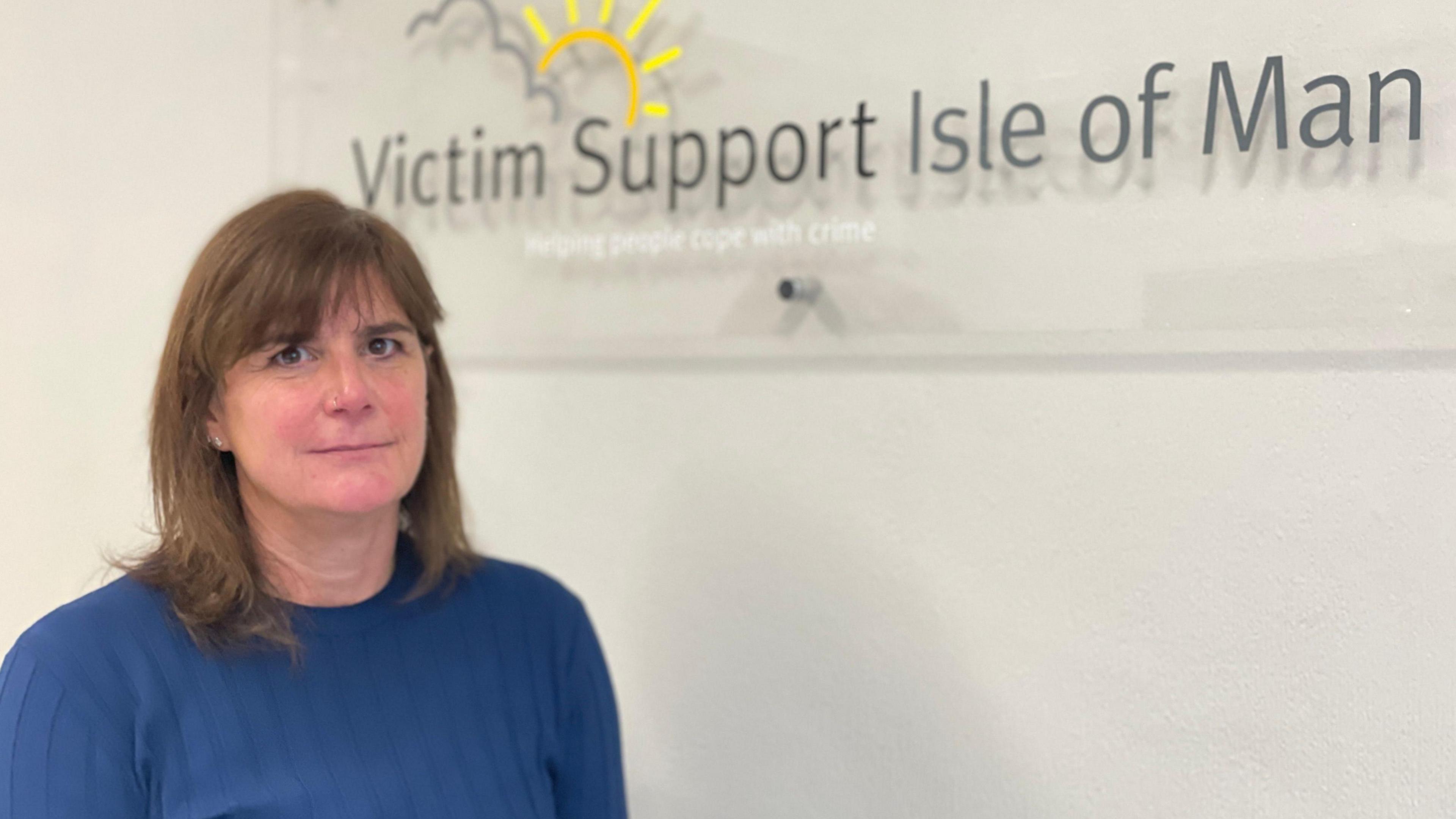 Lorna Trevethan, who has short brown hair and is wearing a blue jumper. She is standing in front of a sign that reads Victim Support Isle of Man.