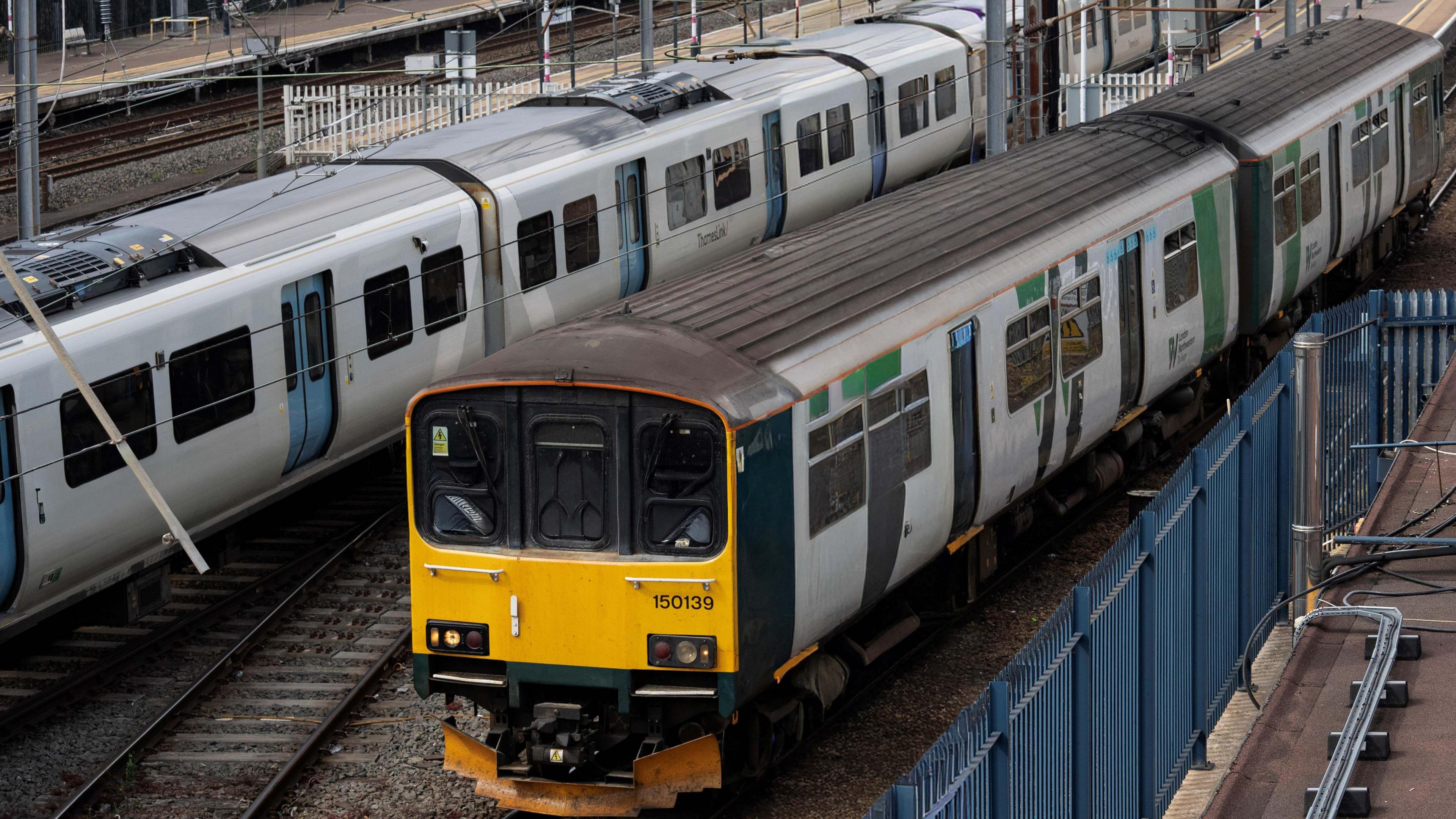 Two trains in Bedford railway station, showing one for the Marston Vale line. There are power cables, tracks a yellow train carriage. Metal fences, to the right and platforms to the right. 