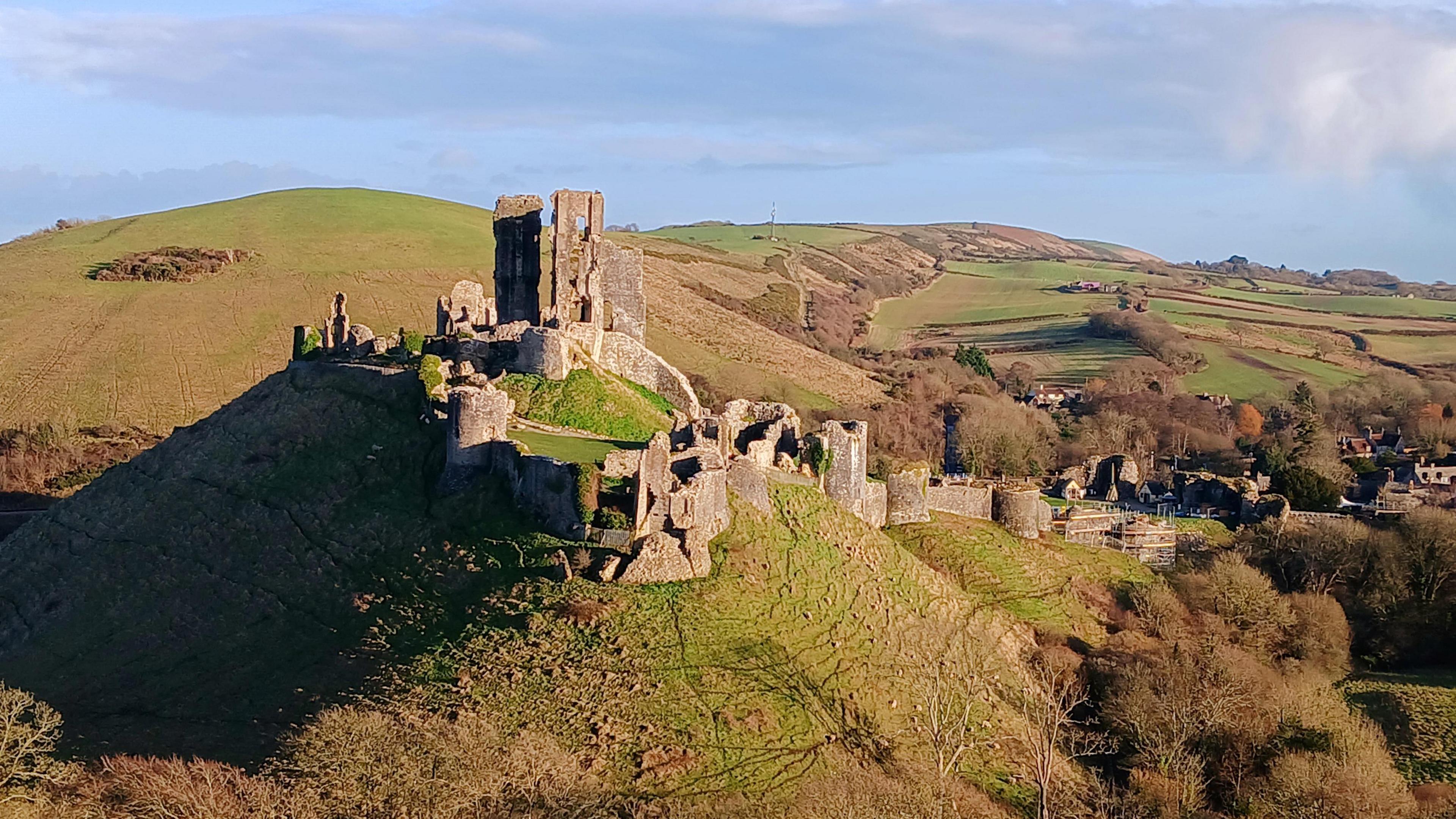 The ruins of Corfe Castle looking regal in this photograph.  The stone castle stands on top of a grassy hill. Behind you can see the rolling hills of the Dorset countryside. It is a bright sunny day with blue sky.