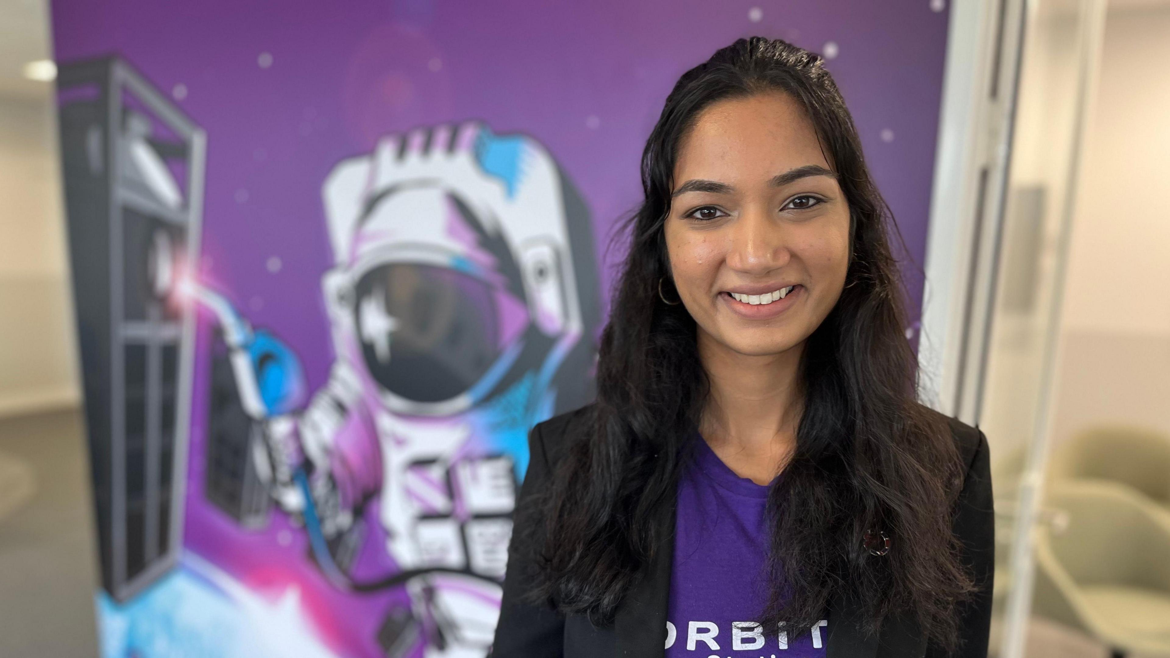 Jas Tiruvuru is wearing a black blazer over a purple shirt with the word 'Orbit' visible in white lettering. Behind her is a graphic image of an astronaut holding a fuel nozzle.