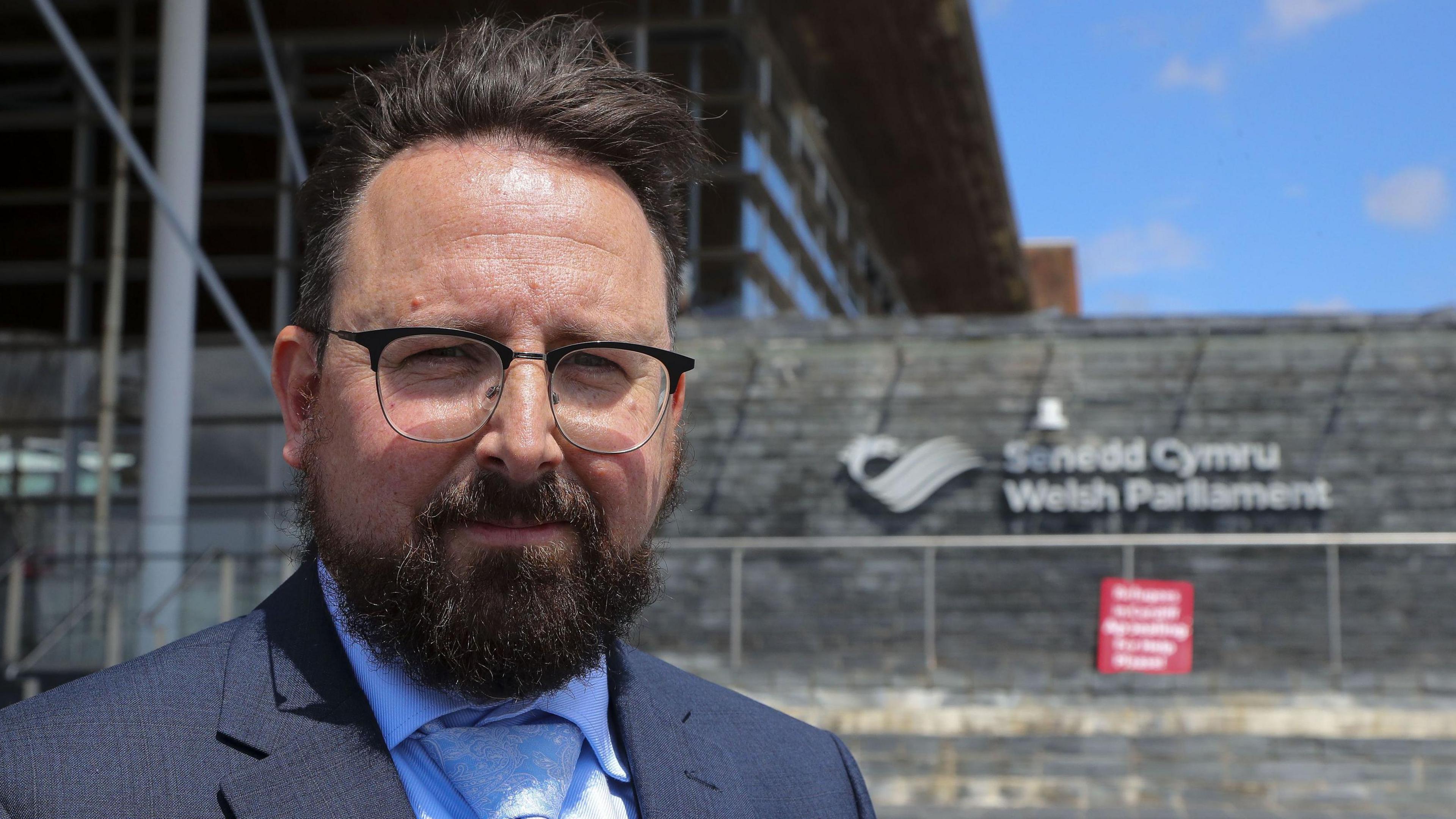 Photograph of Hefin David outside the Senedd. He is wearing a navy suit, blue shirt and tie, dark-rimmed glasses and has a thick dark beard.