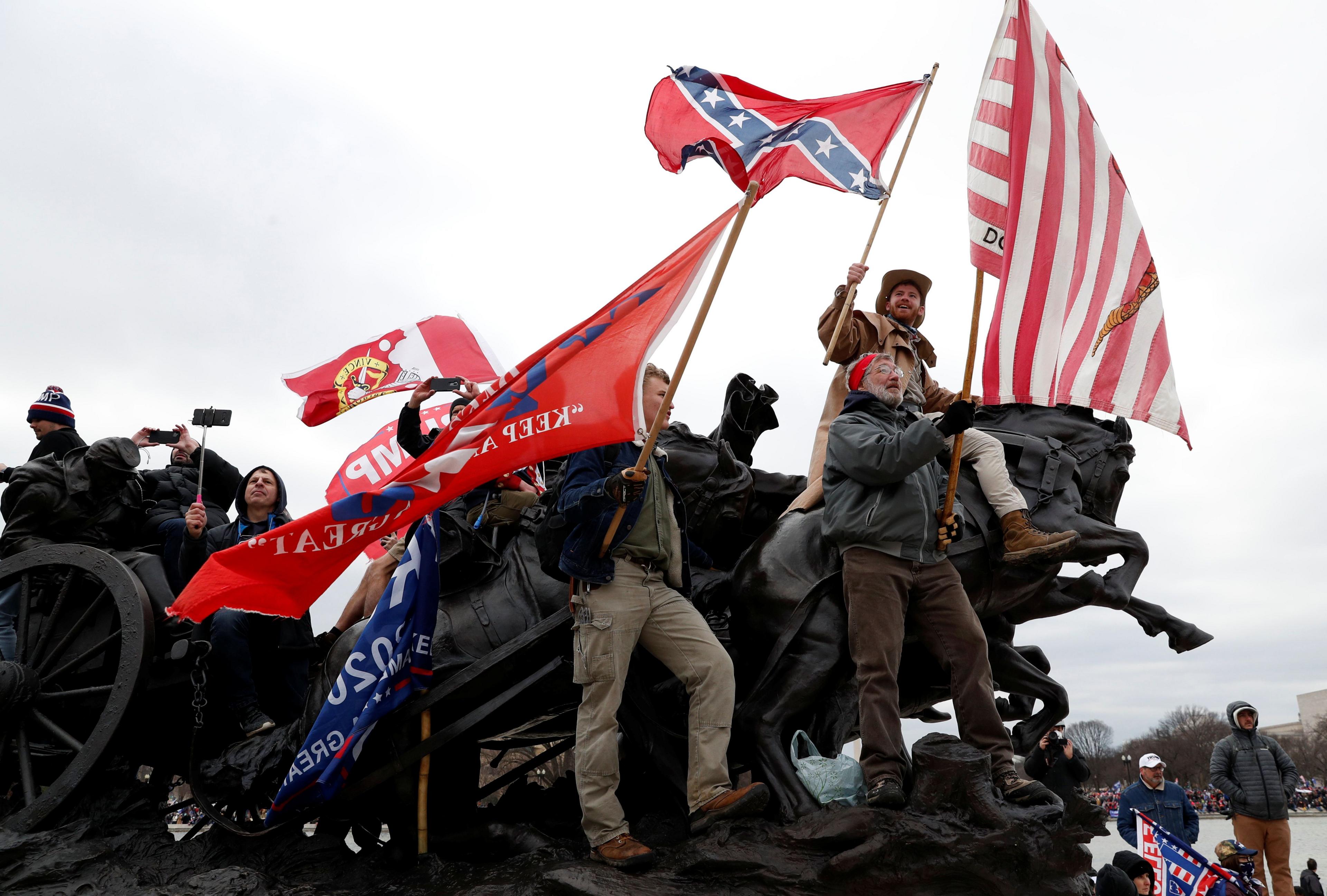 Men atop a statue holding flags saying "Trump" and a Confederate flag