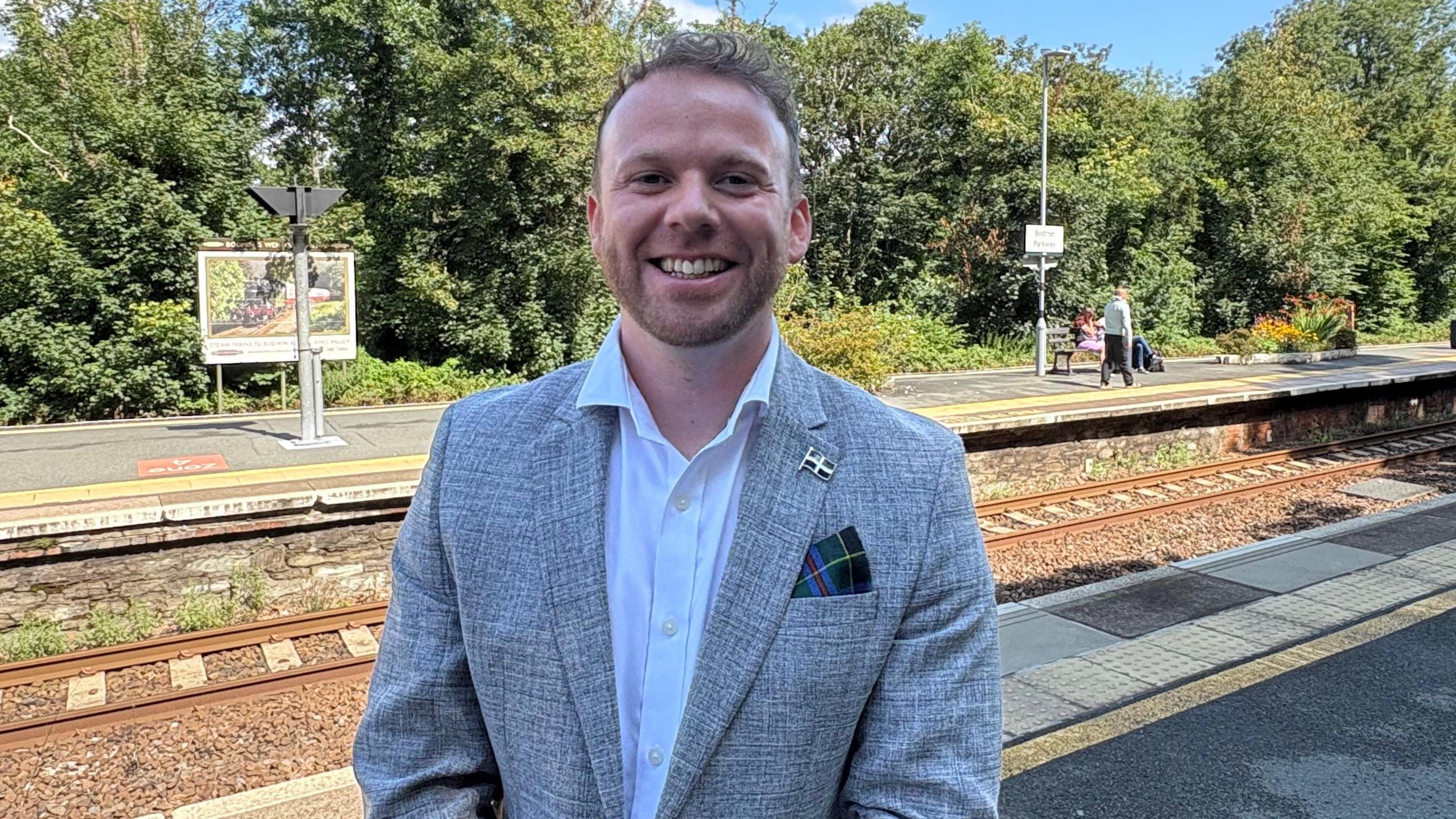 A man with dark blond hair and stubble wearing a white shirt and grey jacket standing on the platform at Bodmin Parkway station with the train tracks and second platform behind him. 