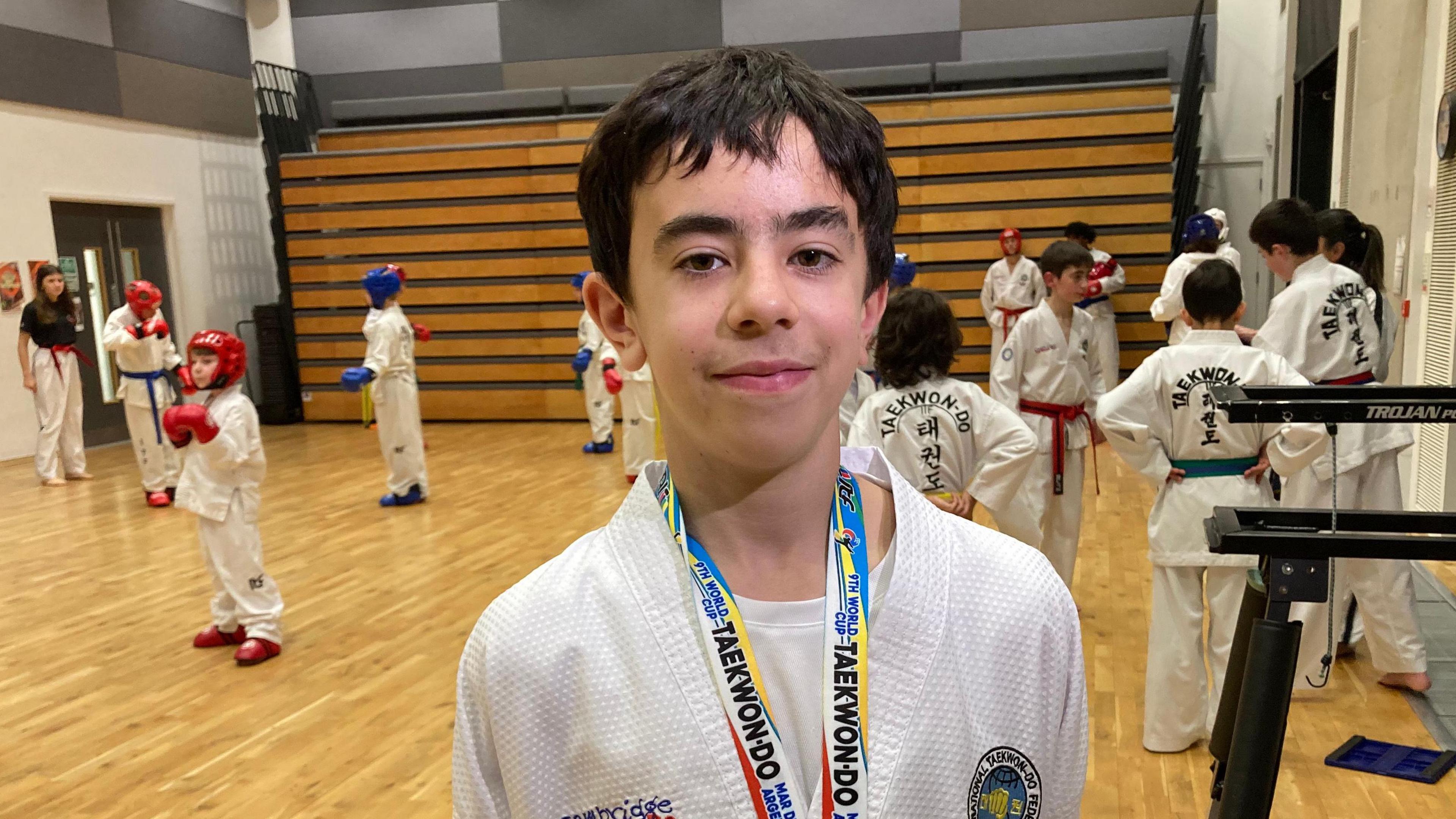 Danyal Kadioglu, 12, with teammates at training. He is standing in a gym with a wooden floor. He is wearing a white martial arts suit and has a medal around his neck. Several young people in white martial arts suits can be seen behind him. Some are wearing boxing gloves and wearing head protectors.